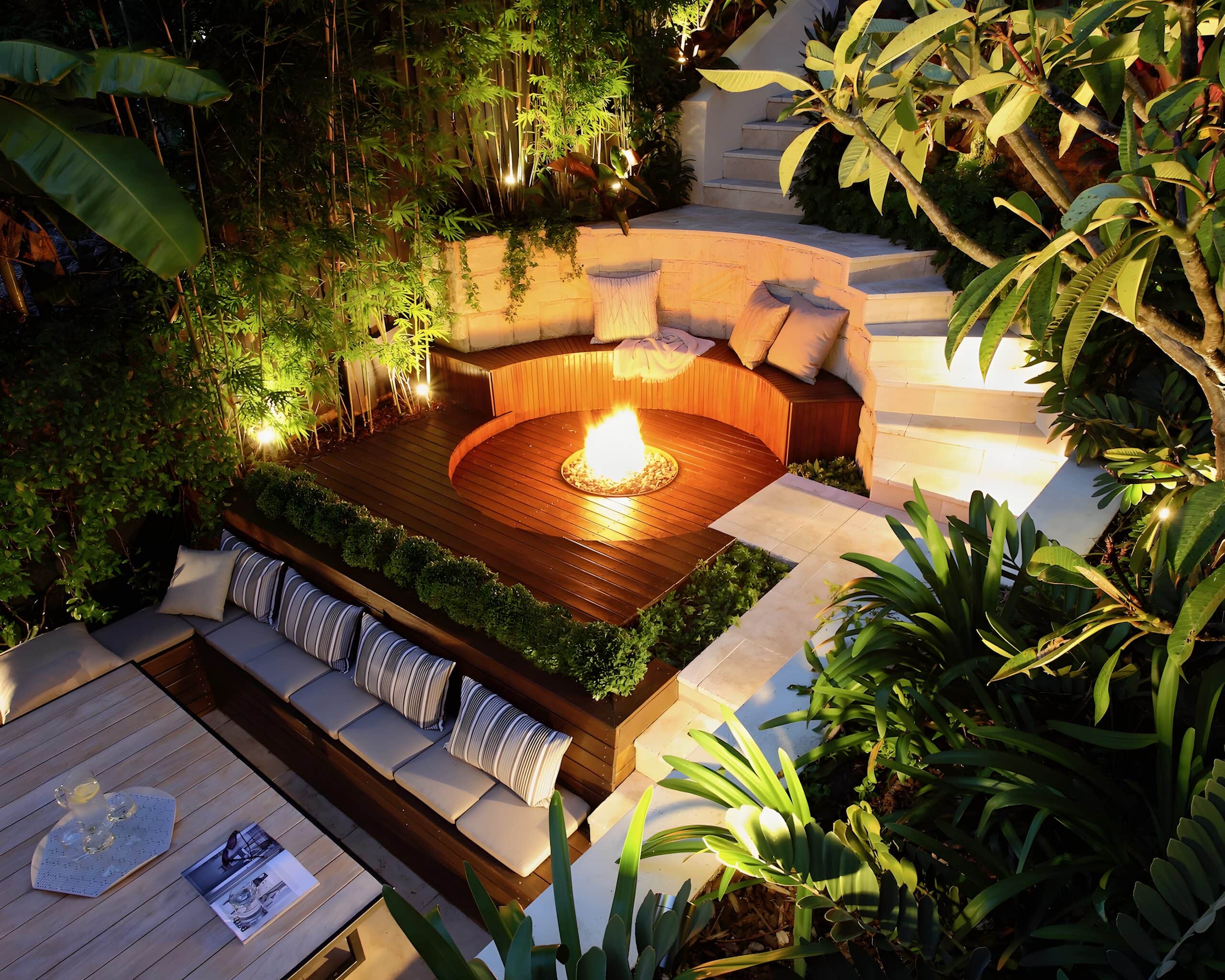 Elevated dusk view of a sunken timber deck with a lit gas fire pit and curved sandstone bench glowing under garden lighting