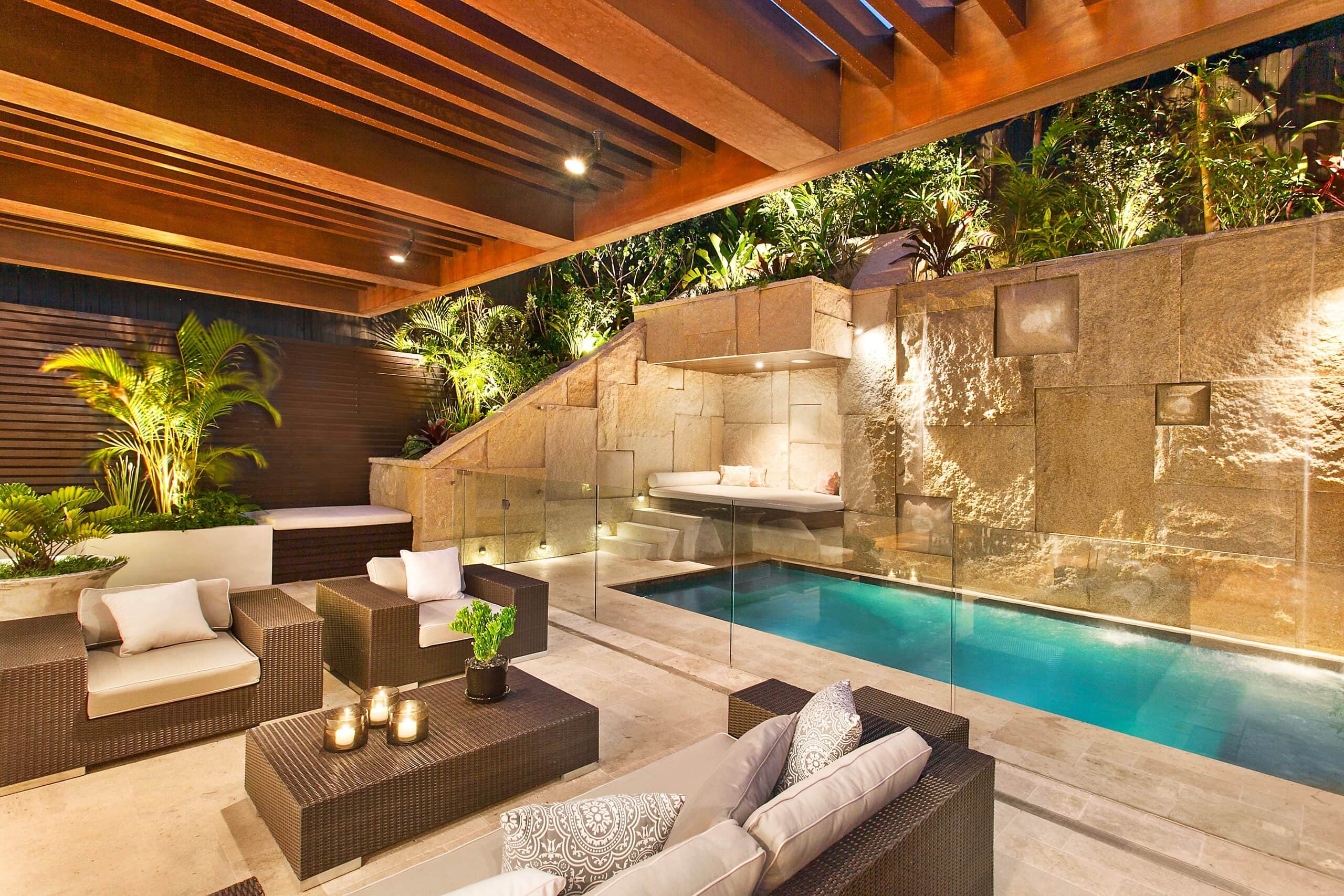 Coastal courtyard at dusk with a plunge pool glowing beside a sandstone waterfall wall under a warmly lit timber pergola with wicker lounges and candles