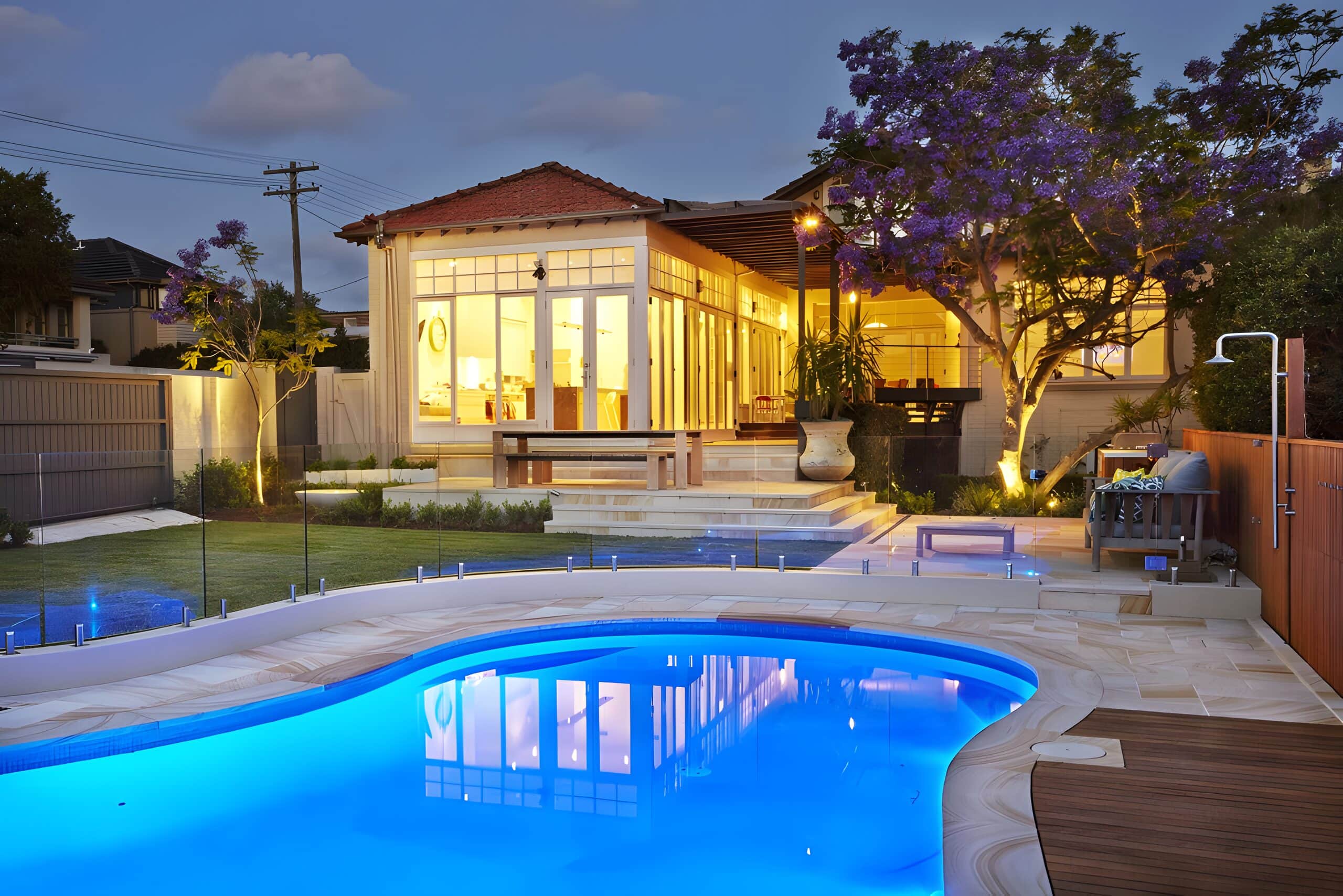 Kidney-shaped pool glowing blue at dusk beside sandstone paving and a warmly lit garden room
