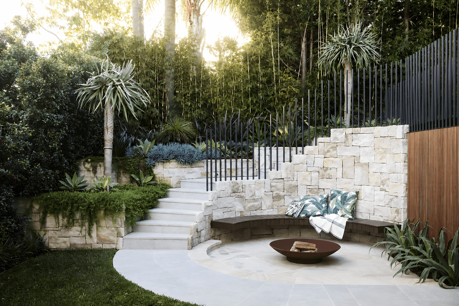 Curved sandstone firepit terrace with stone steps and lush subtropical planting in soft daylight