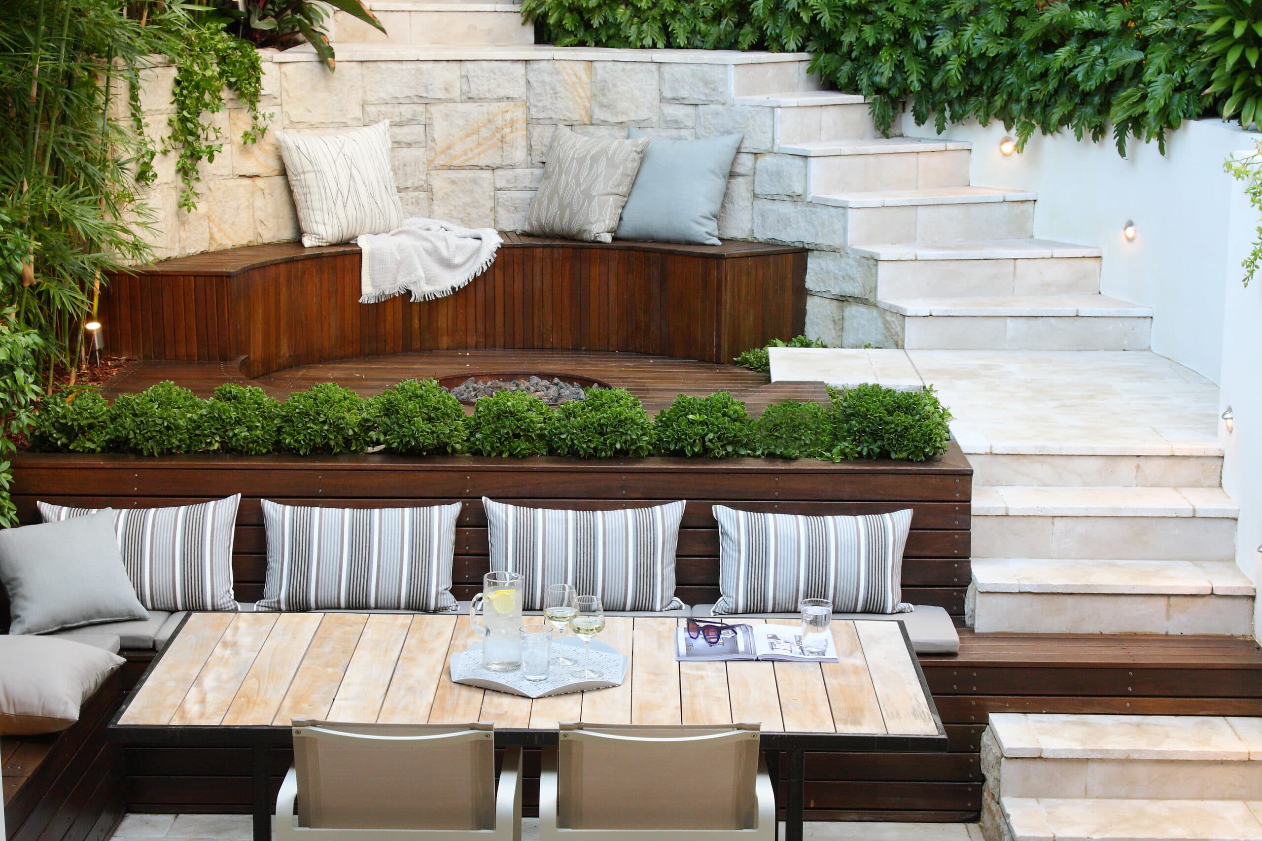Curved timber bench with cushions and a throw against a sandstone wall above a clipped box hedge and fire pit