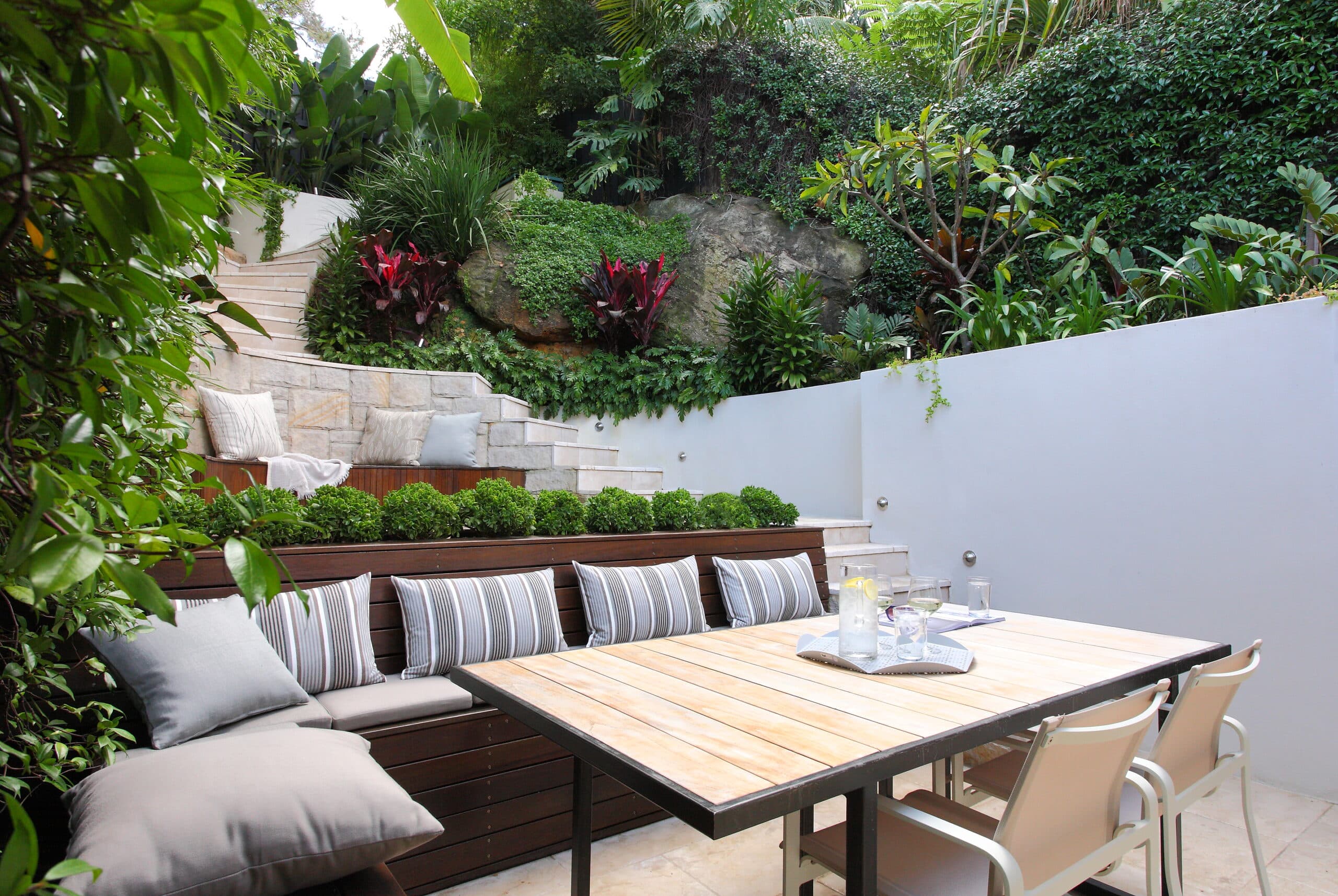 Sunlit banquette and teak dining table below a sandstone retaining wall with a moss covered boulder and red cordylines