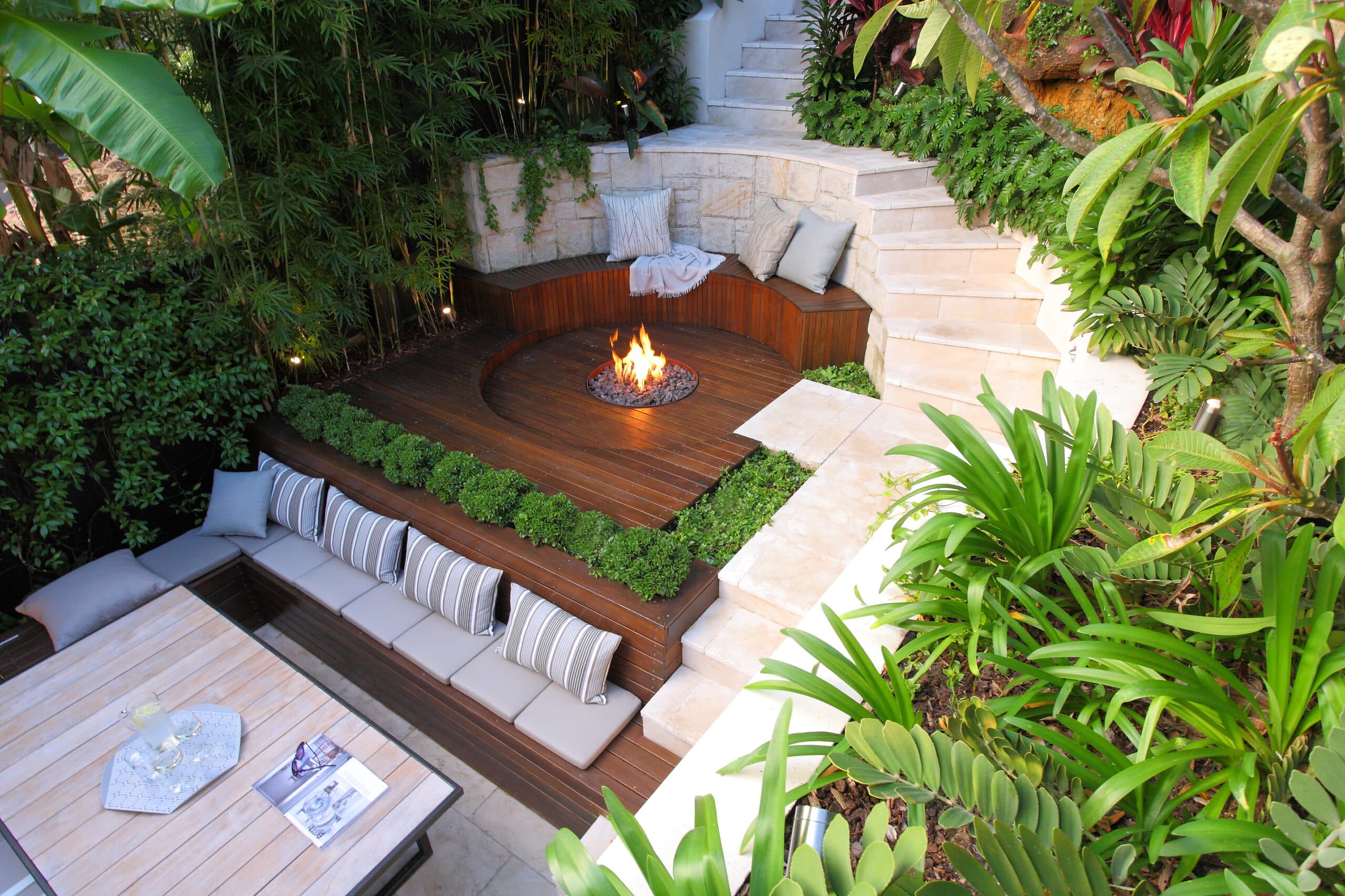 Looking down onto a burning fire pit ringed by a curved timber bench with frangipani and agapanthus alongside pale stone steps