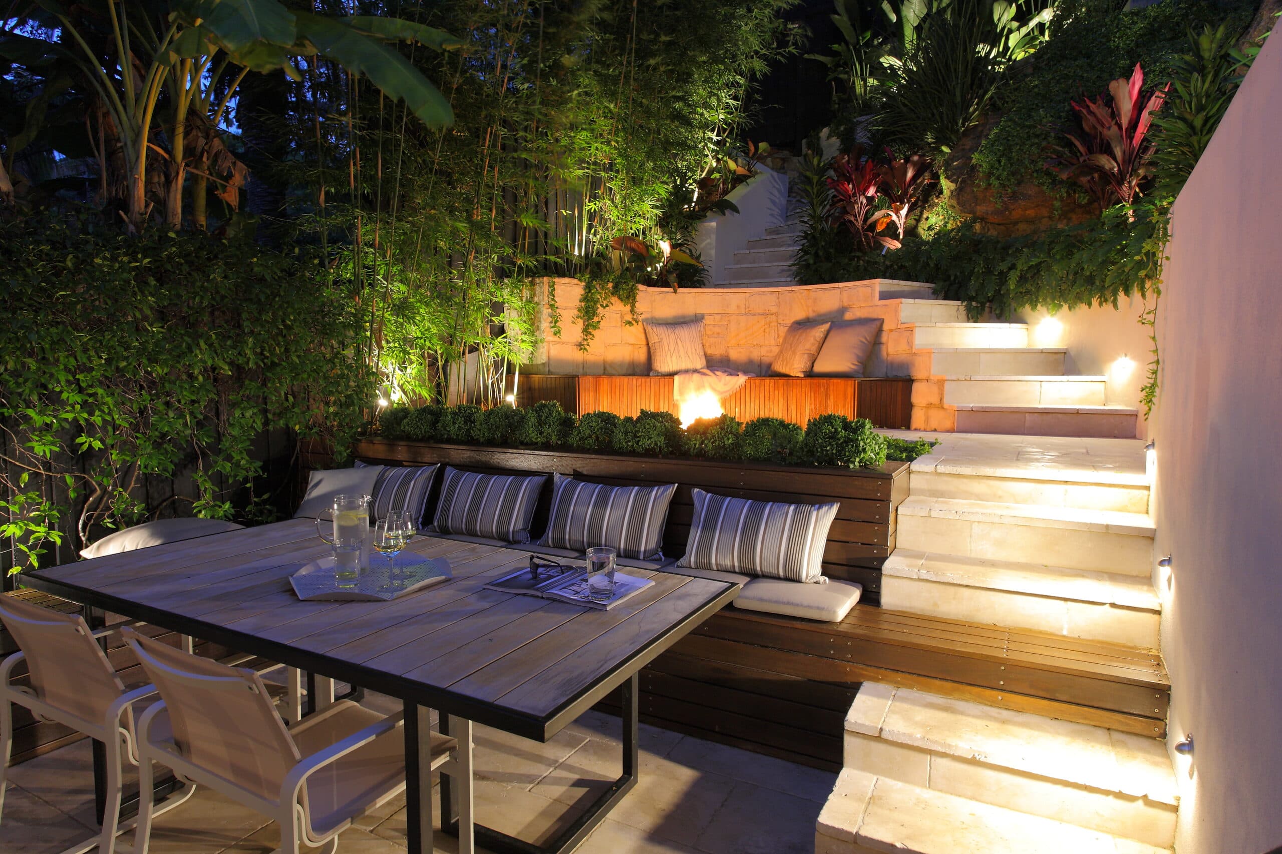 Evening dining setting with warm uplighting on bamboo and cordylines beside sandstone steps and a distant fire glow