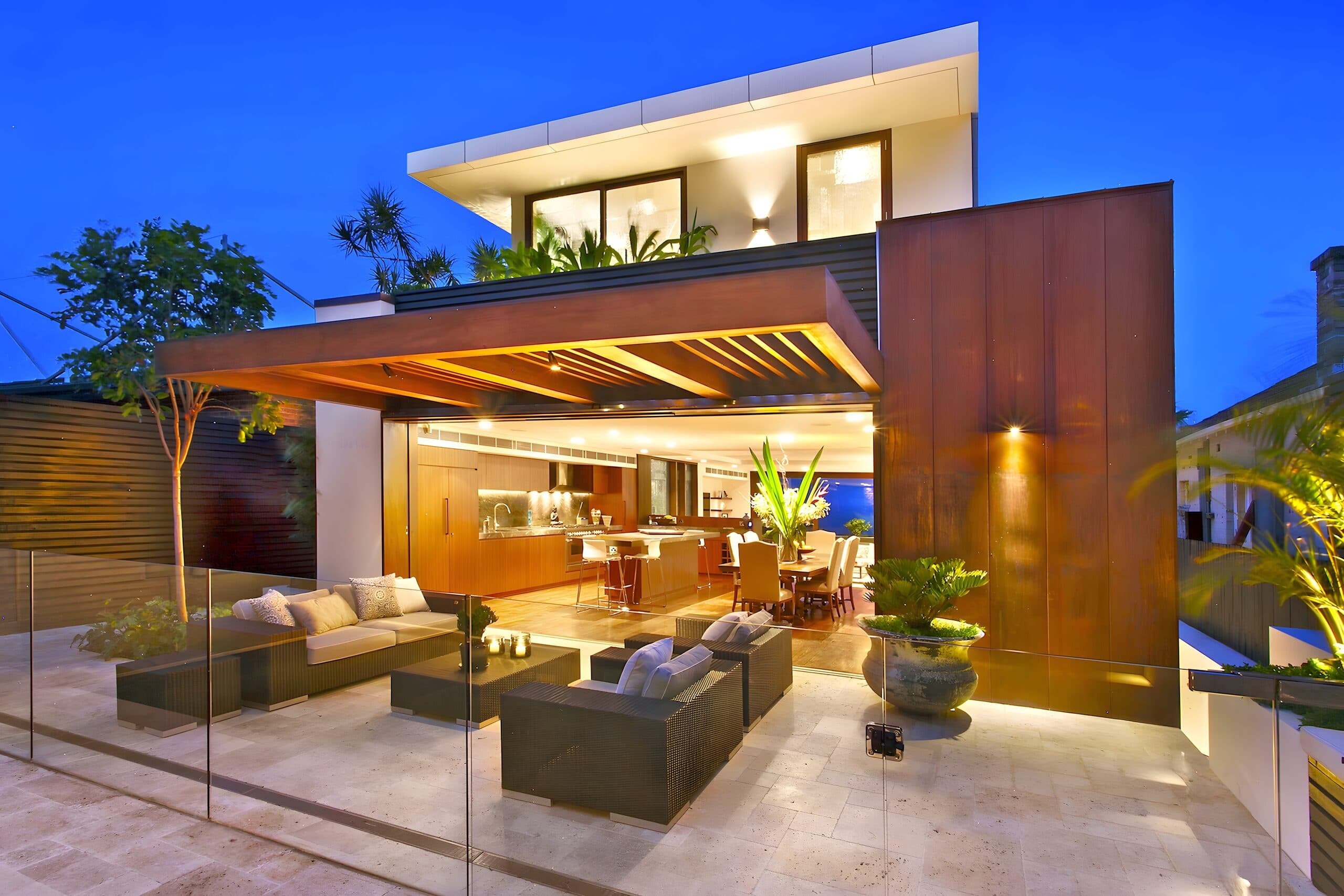 Modern two-storey home lit from within at dusk opening onto a travertine terrace behind glass balustrade