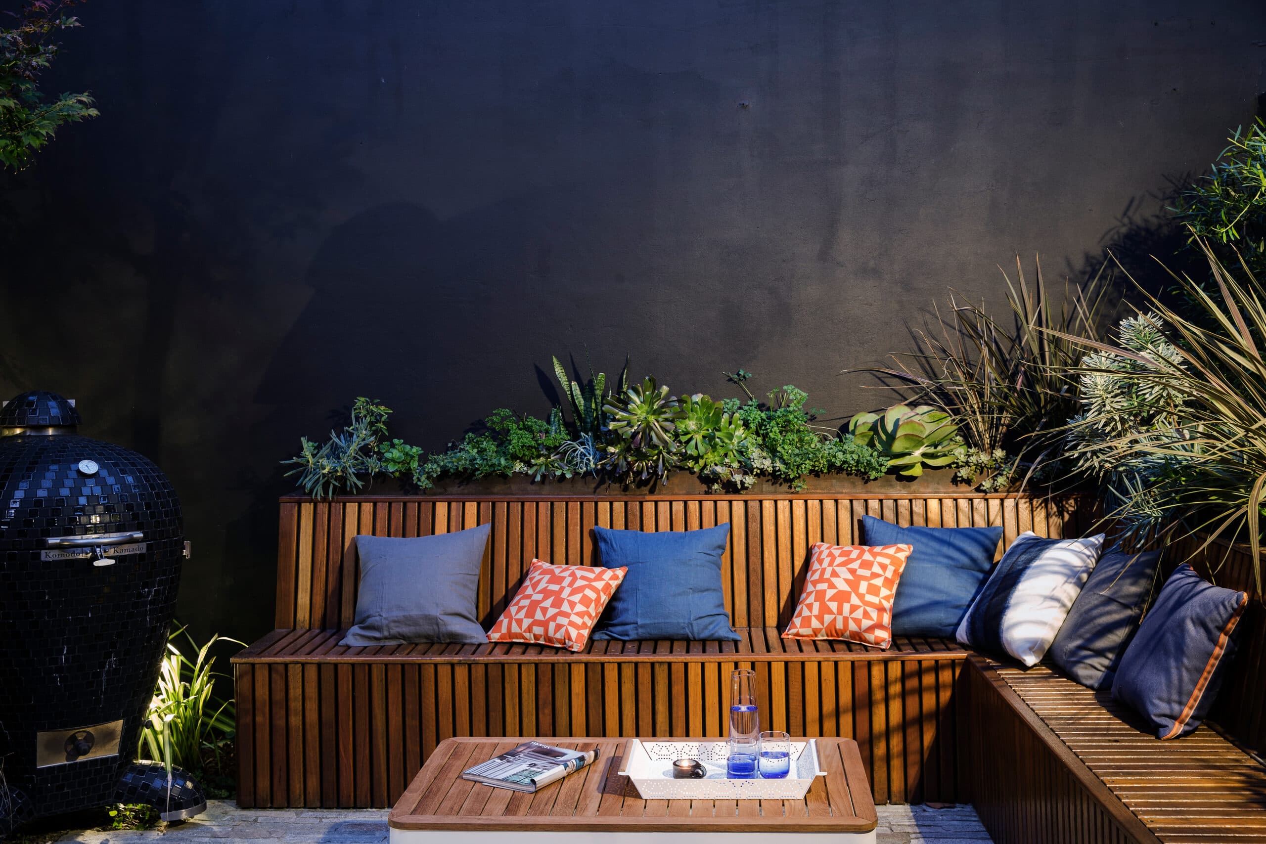 Slatted bench and lit kamado grill beneath a planted charcoal wall at night