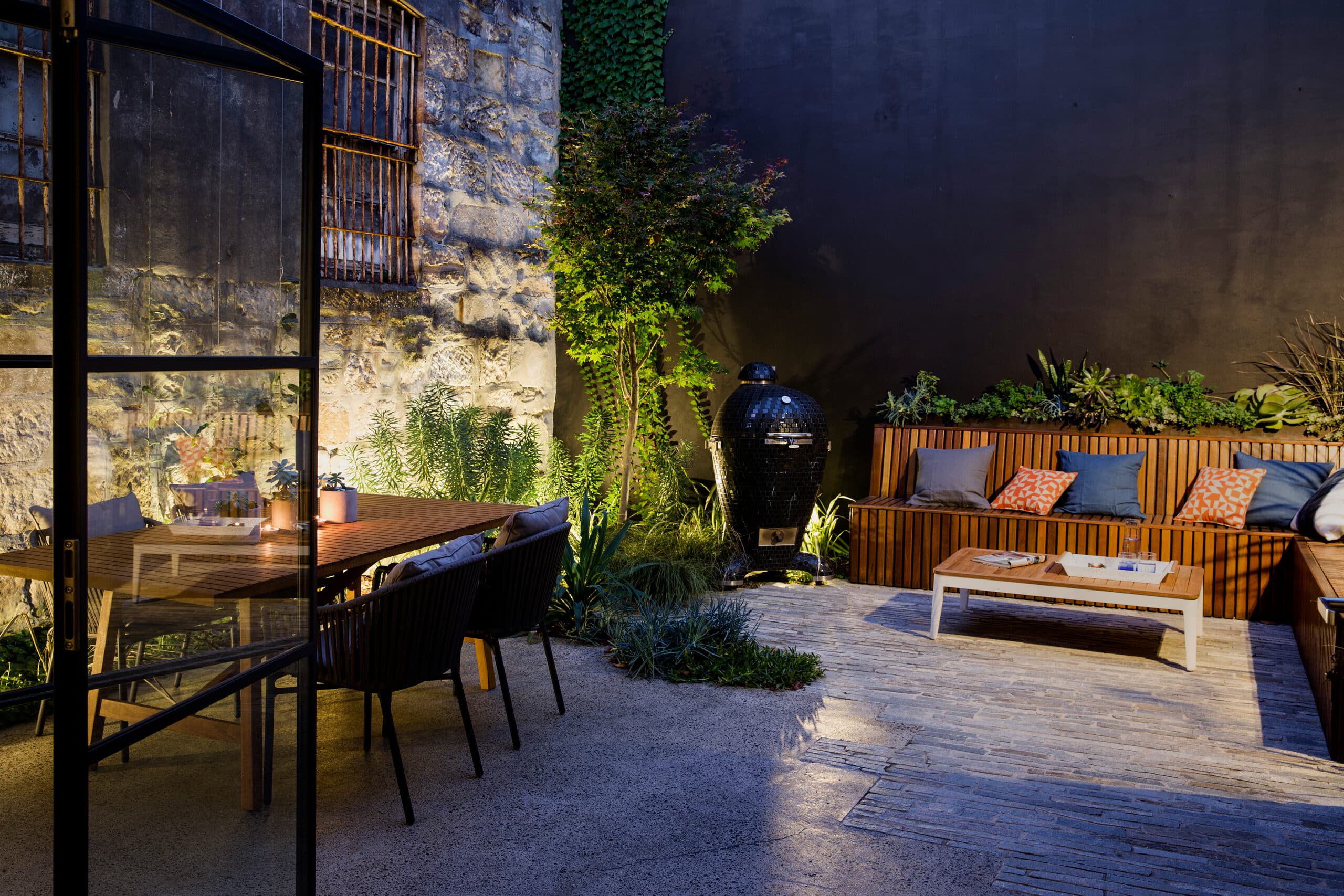 Evening courtyard lit against the heritage sandstone wall with a maple and kamado grill
