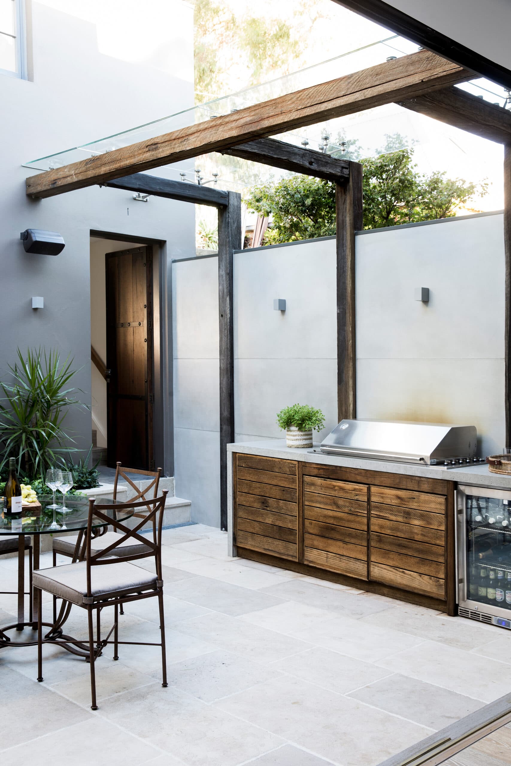 Outdoor kitchen with reclaimed timber cabinetry and stainless barbecue beside a glass dining setting