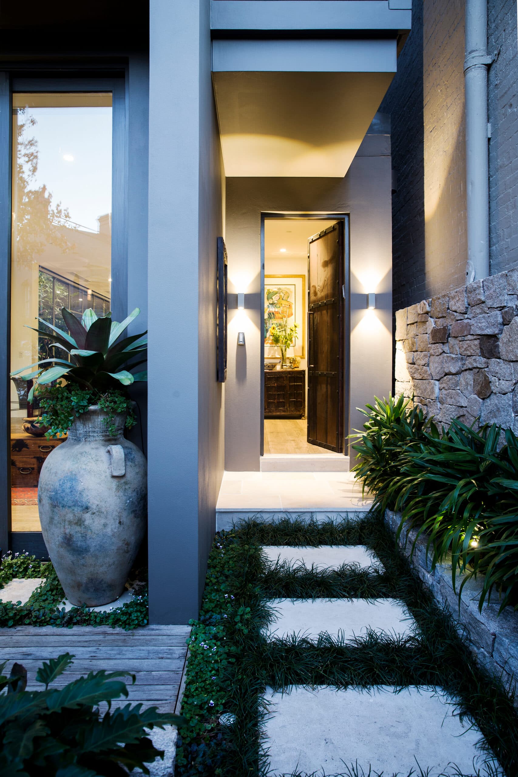 Open timber entry door glowing at dusk between wall lights with an urn and bromeliad