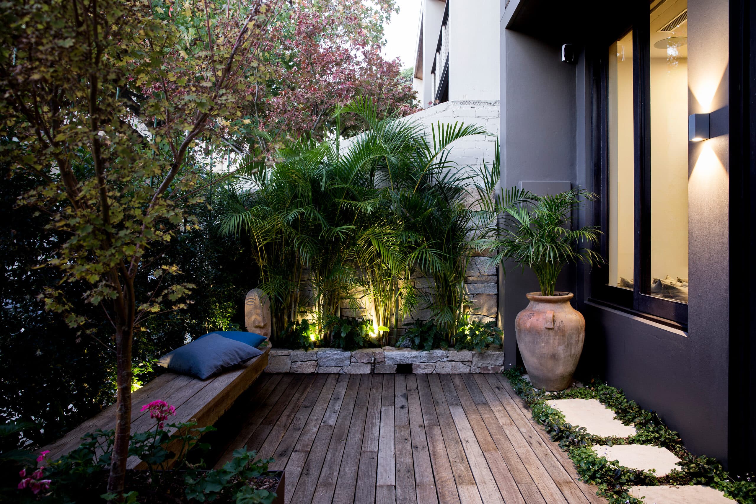 Uplit palms and a stone planter at dusk beside a timber bench and tall terracotta urn
