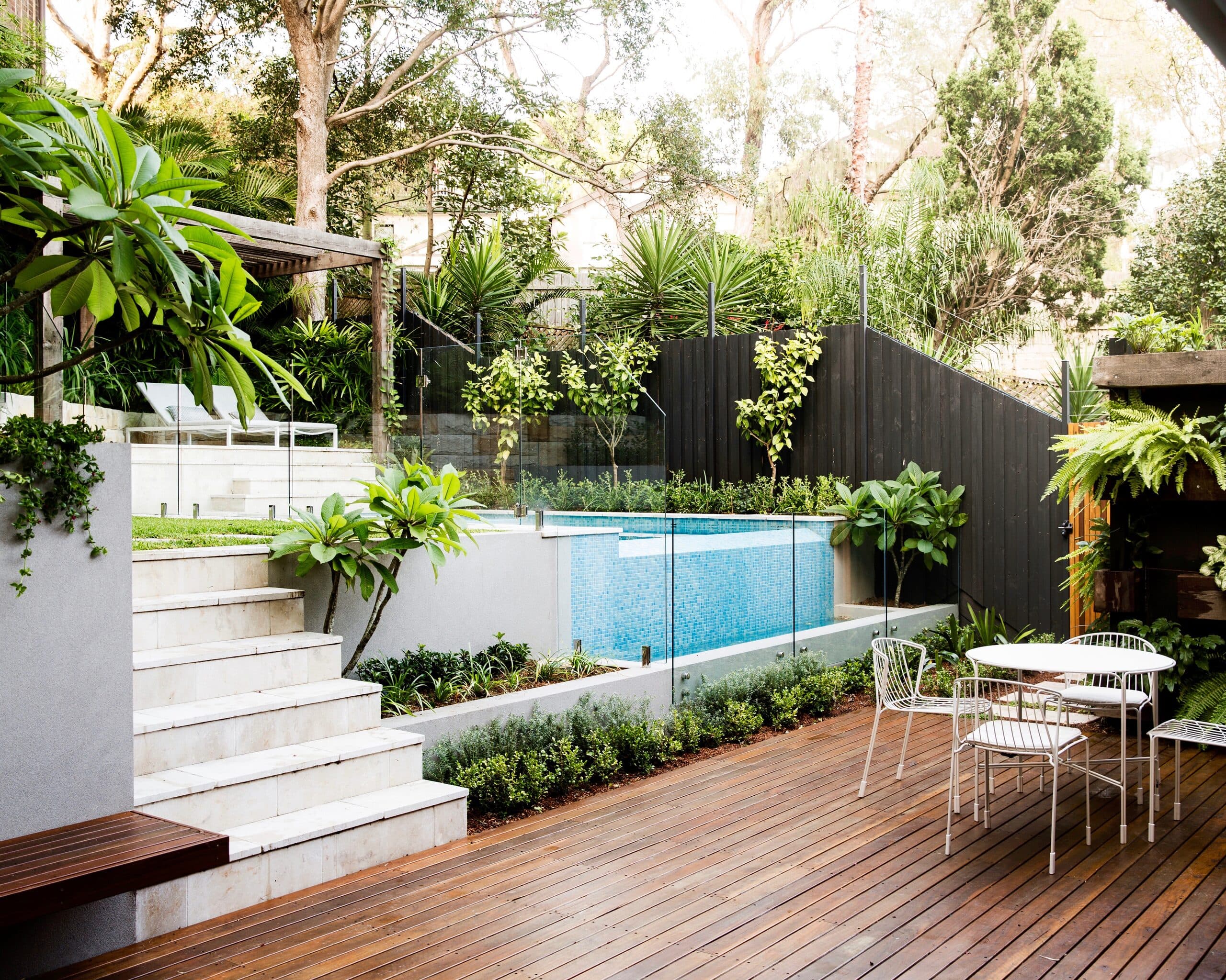 Timber deck and white wire dining set beside a glass fenced plunge pool below terraced sandstone planting