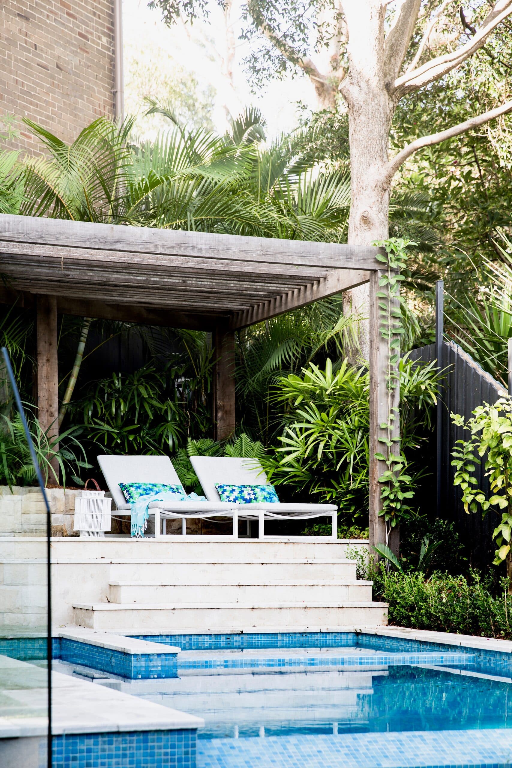 Sun loungers under a timber pergola framed by tropical planting above a mosaic tiled pool