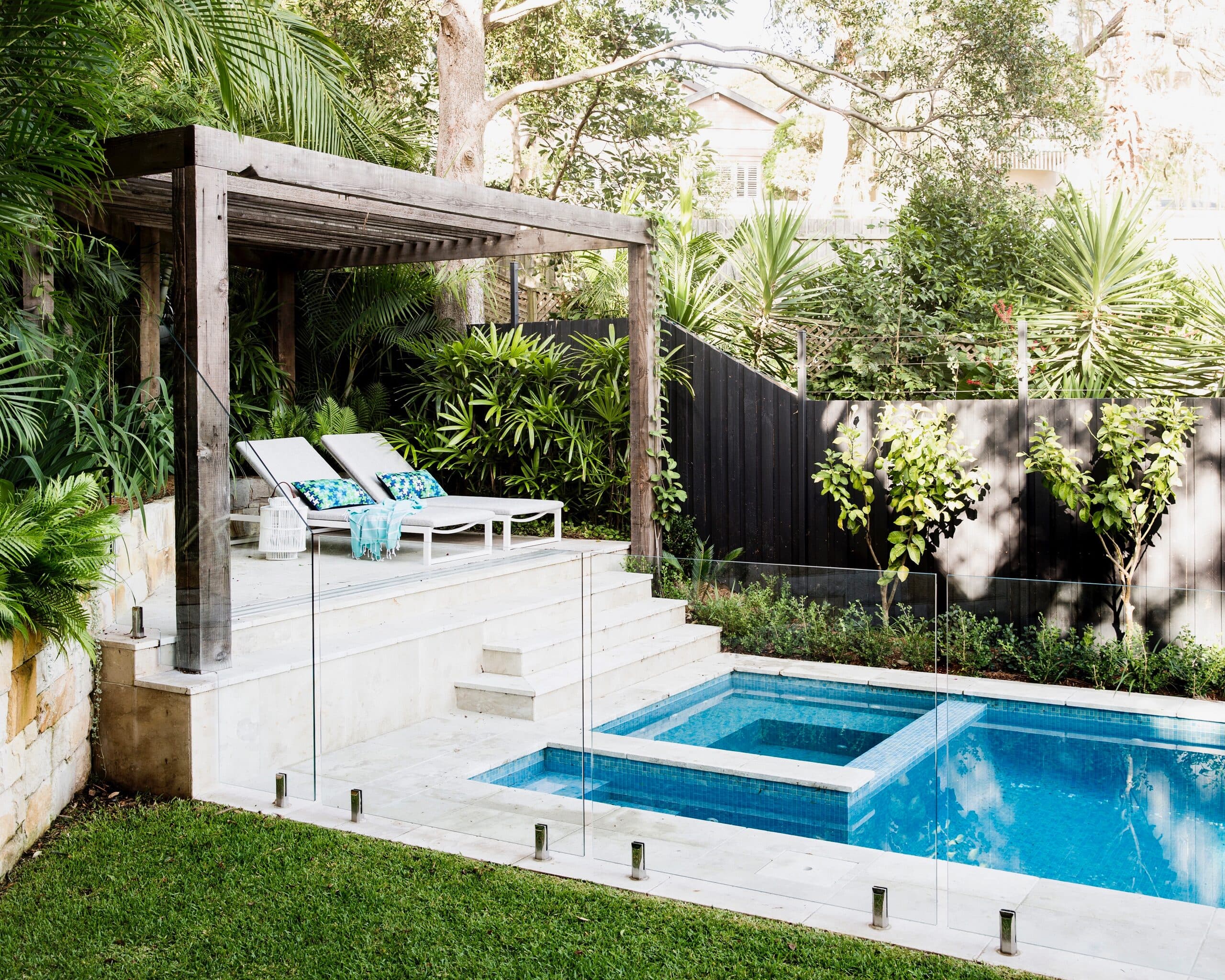 Timber pergola shading loungers on a paved terrace beside a glass fenced pool and spa with lawn
