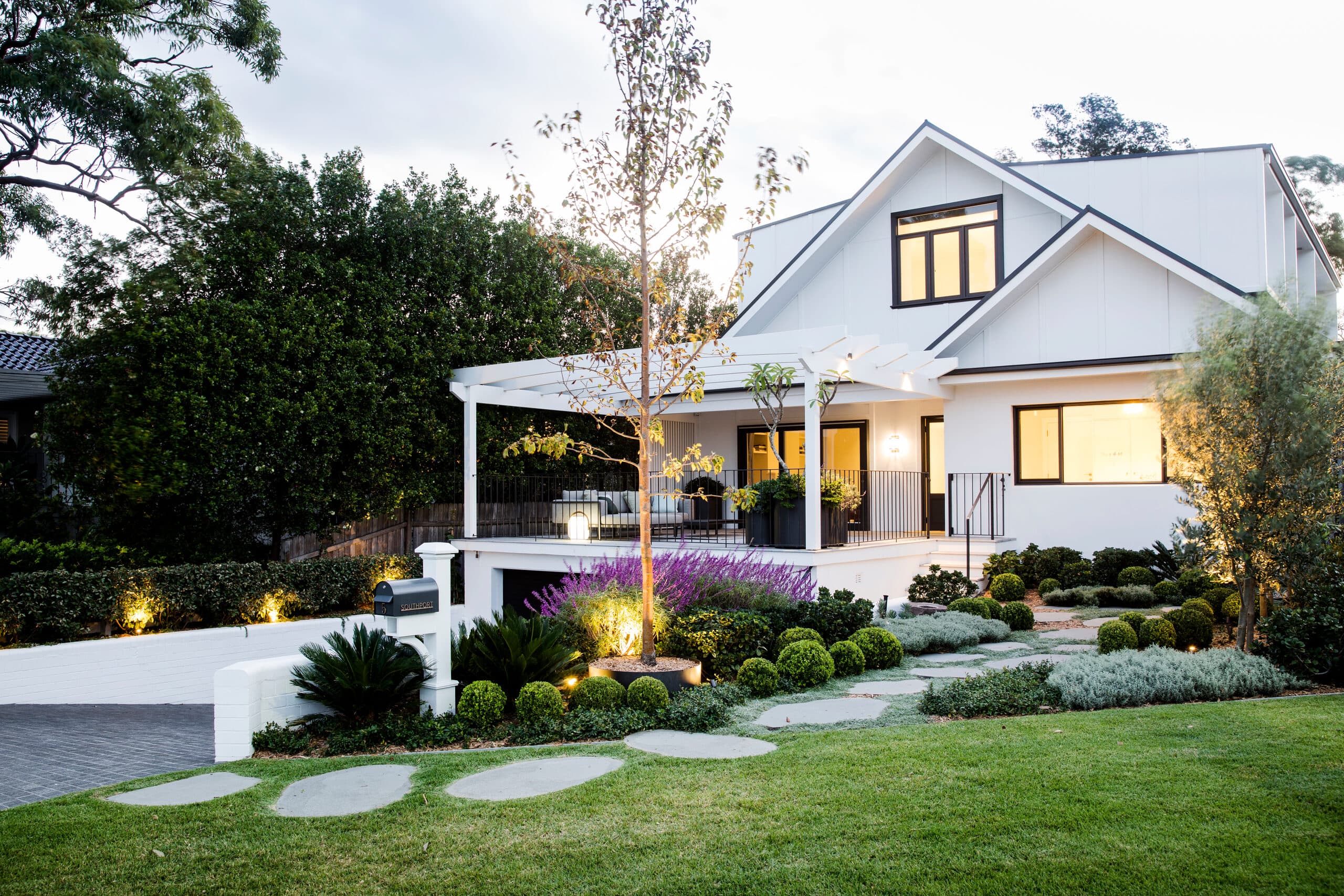 Wide view of the home at dusk with garden lighting, purple salvia and clipped greenery