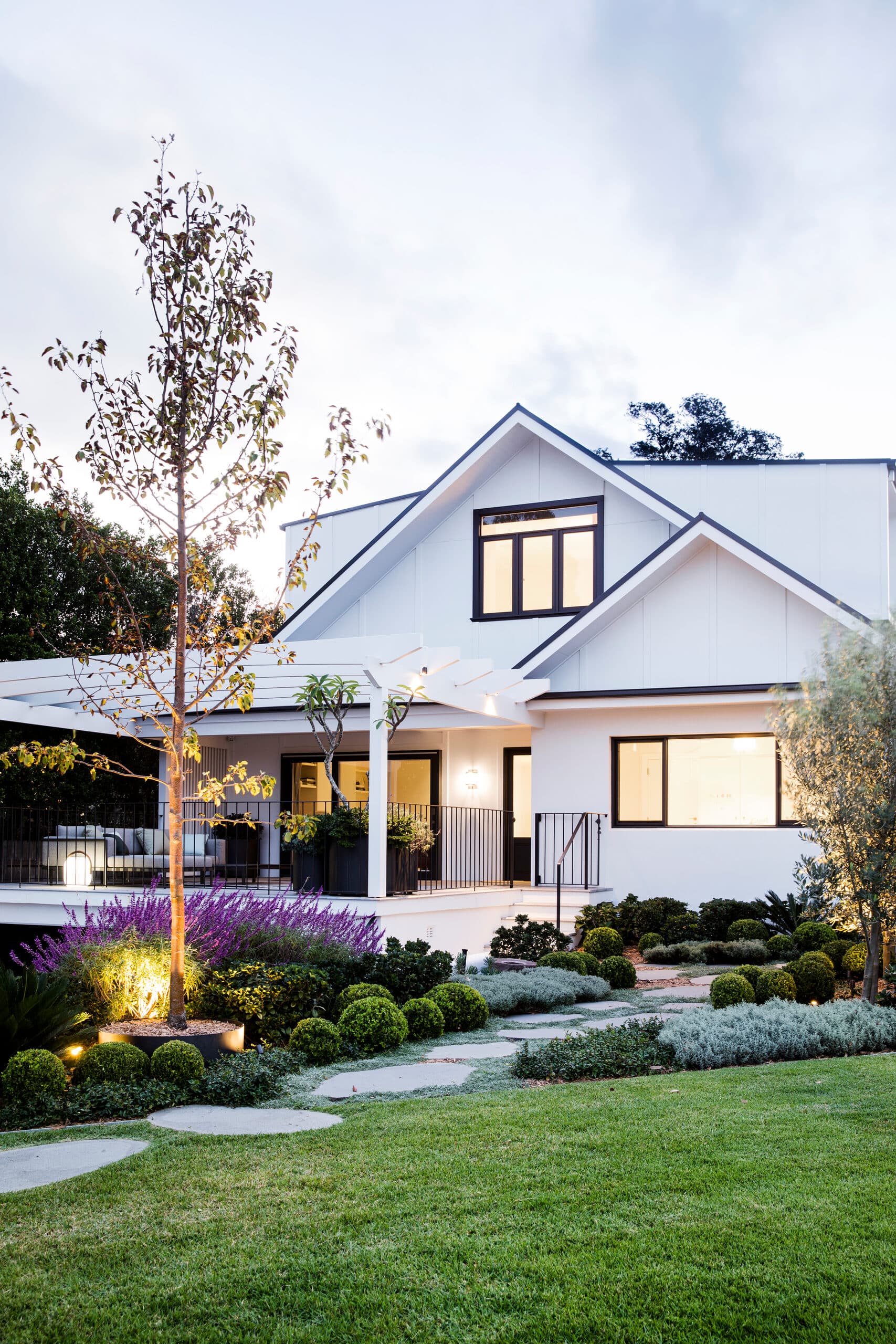 Twin gabled house lit at dusk above uplit salvia, box balls and a stone path