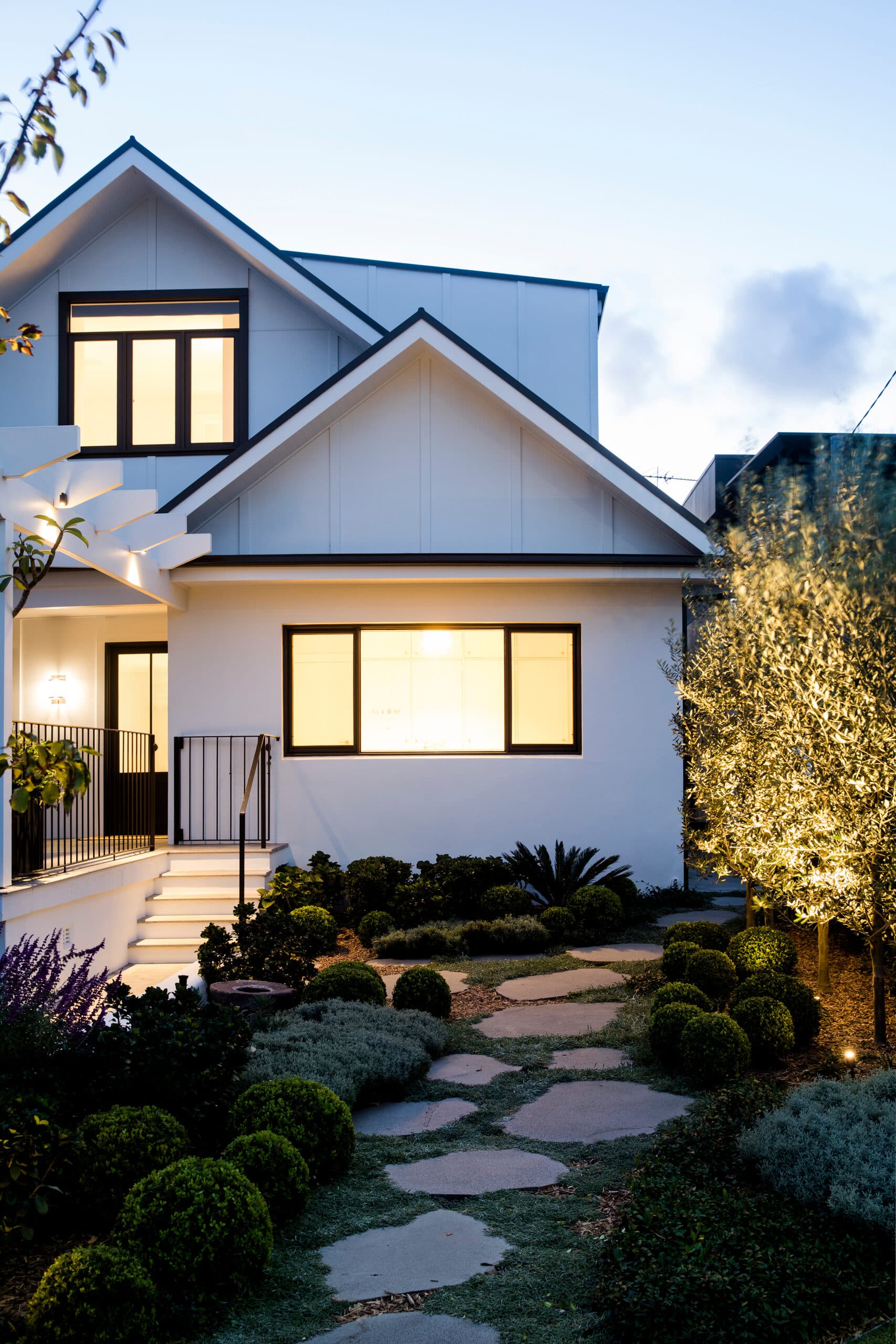Two storey white home glowing at dusk above a lit stepping stone path and topiary