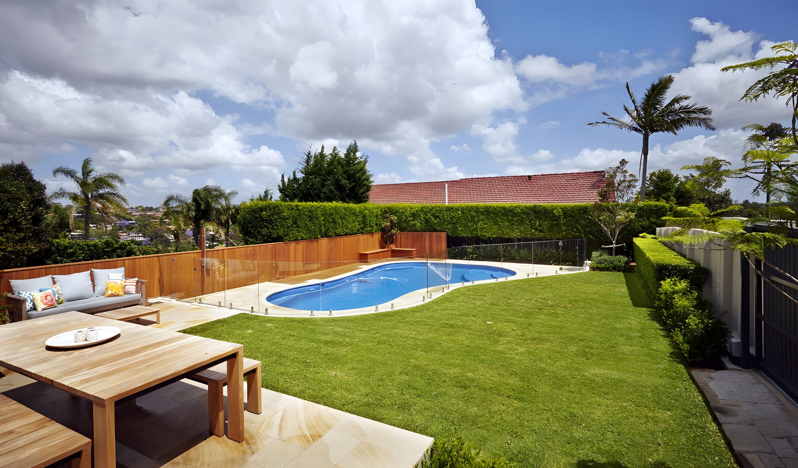 Oval pool and level lawn behind glass fencing with a timber dining terrace and clipped hedges