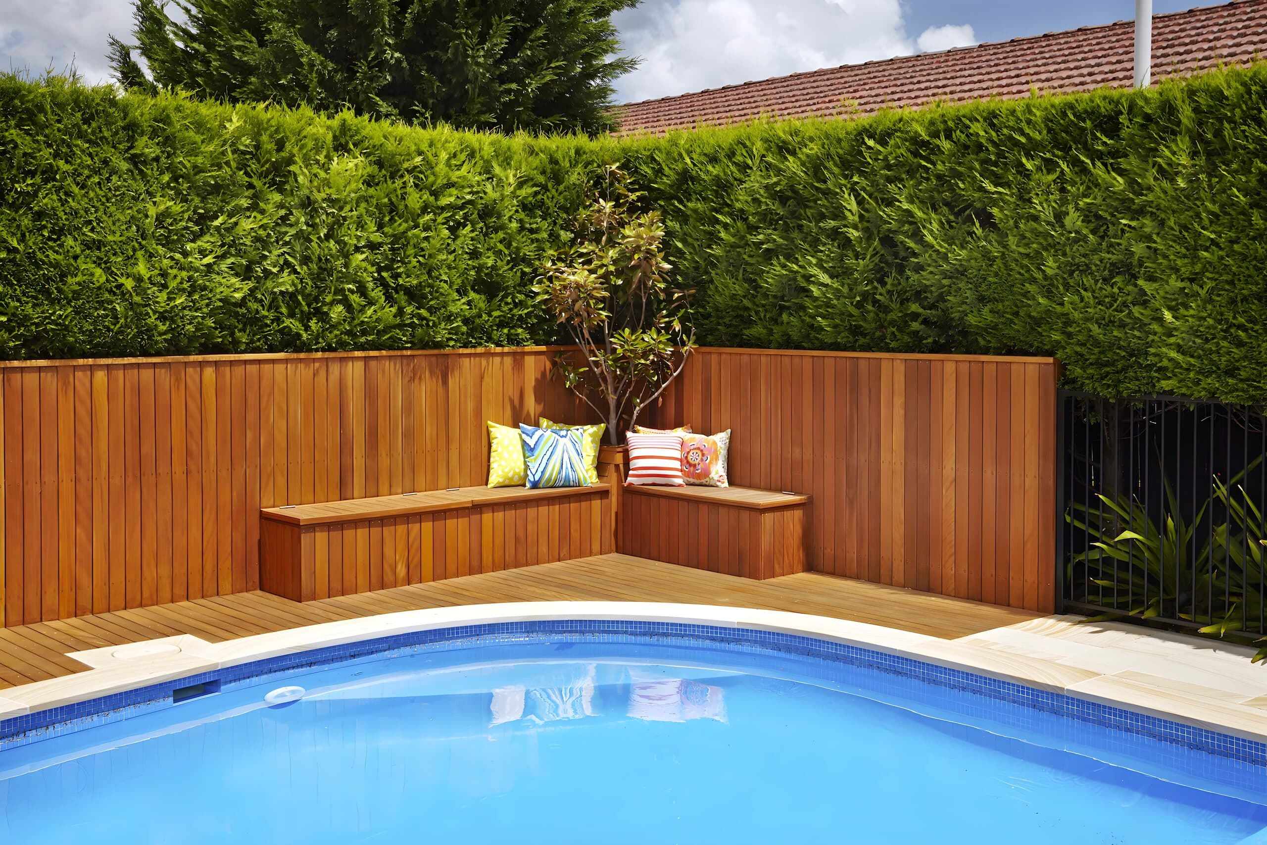 Built-in timber bench with bright cushions tucked into a pool corner below a tall conifer hedge