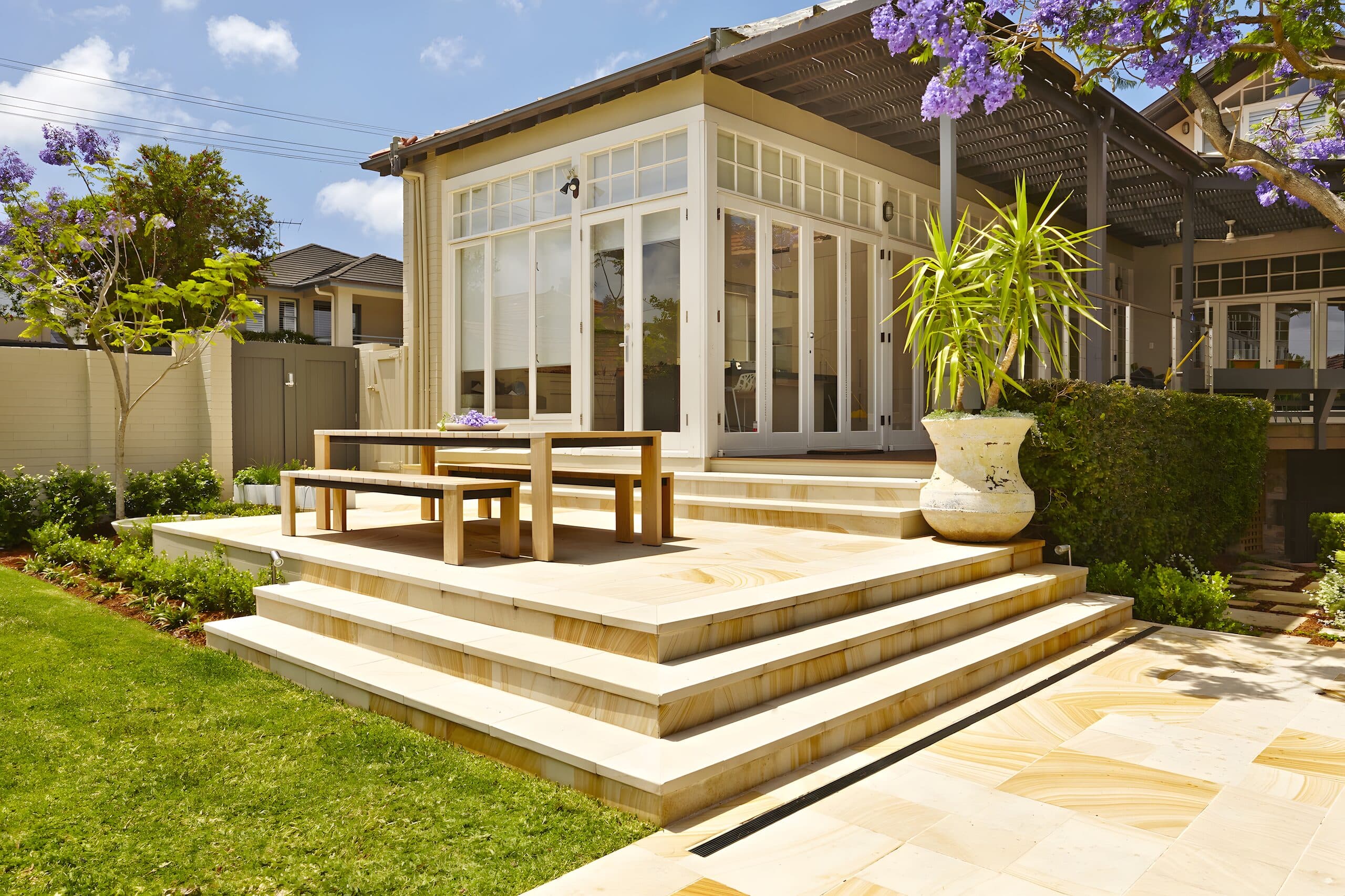 Broad sandstone entertaining terrace with a timber dining setting stepping up to a glass-walled garden room beneath a flowering jacaranda
