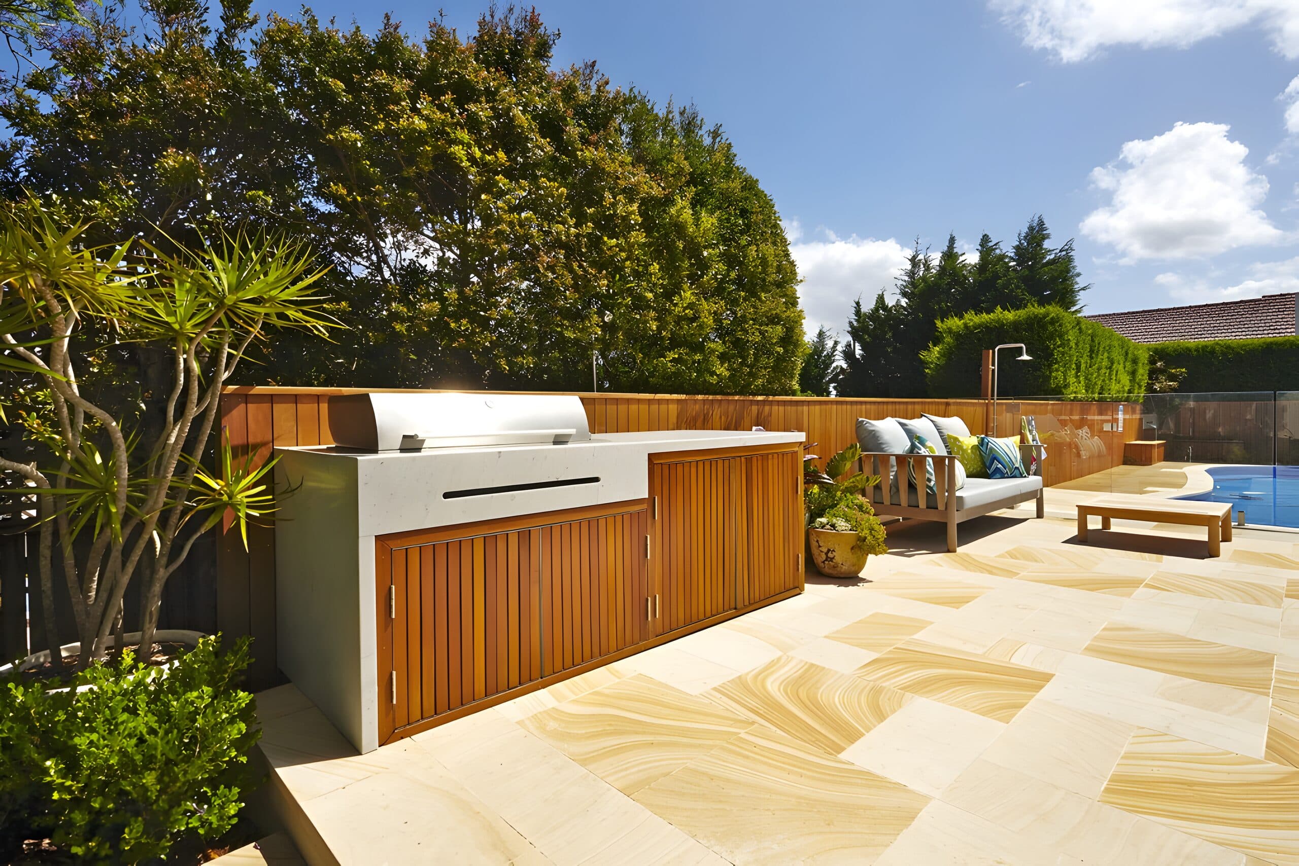 Outdoor kitchen with a hooded barbecue and stone benchtop on sandstone paving beside the pool