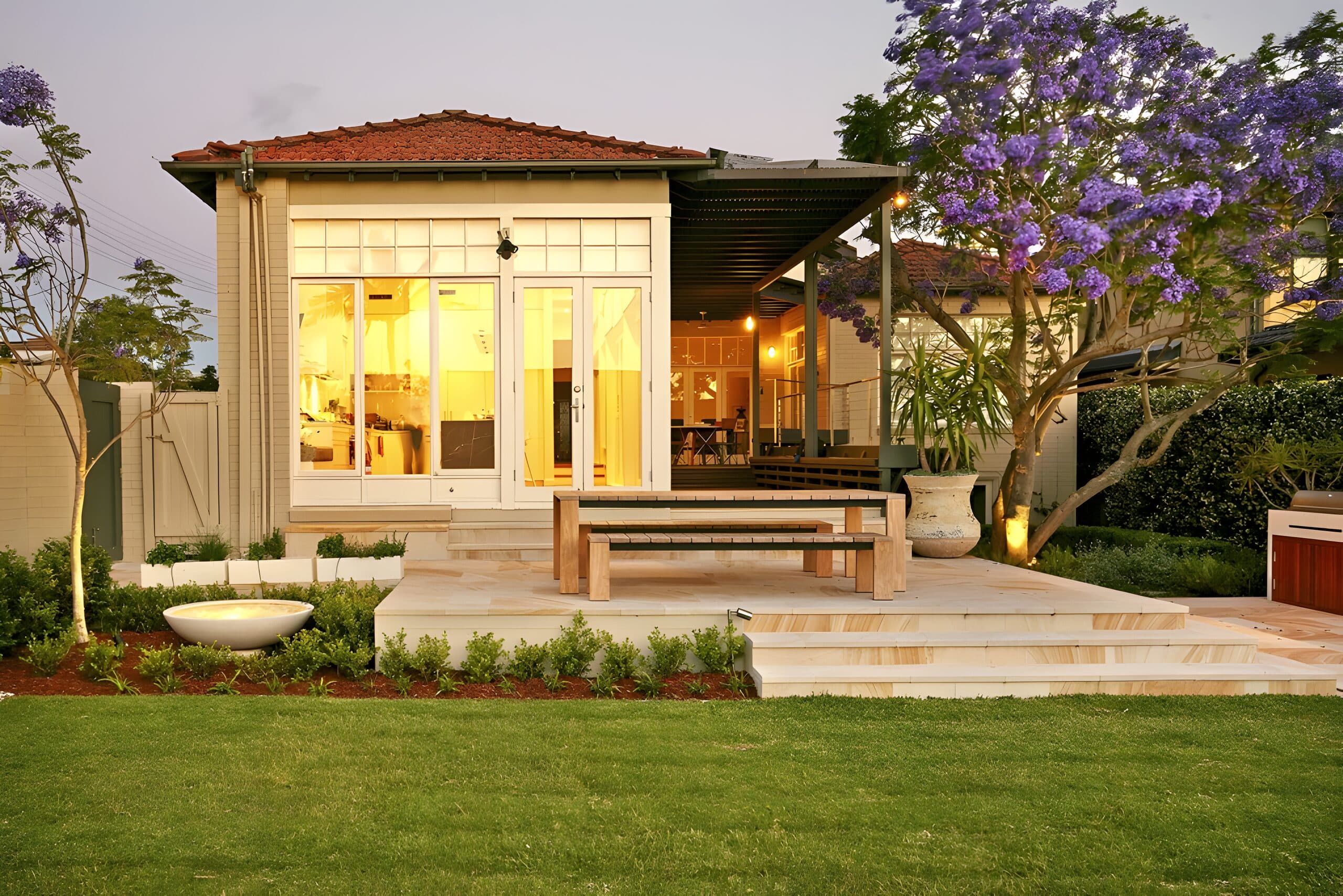Rear of the home lit warmly at dusk with a timber table on the sandstone terrace above the lawn