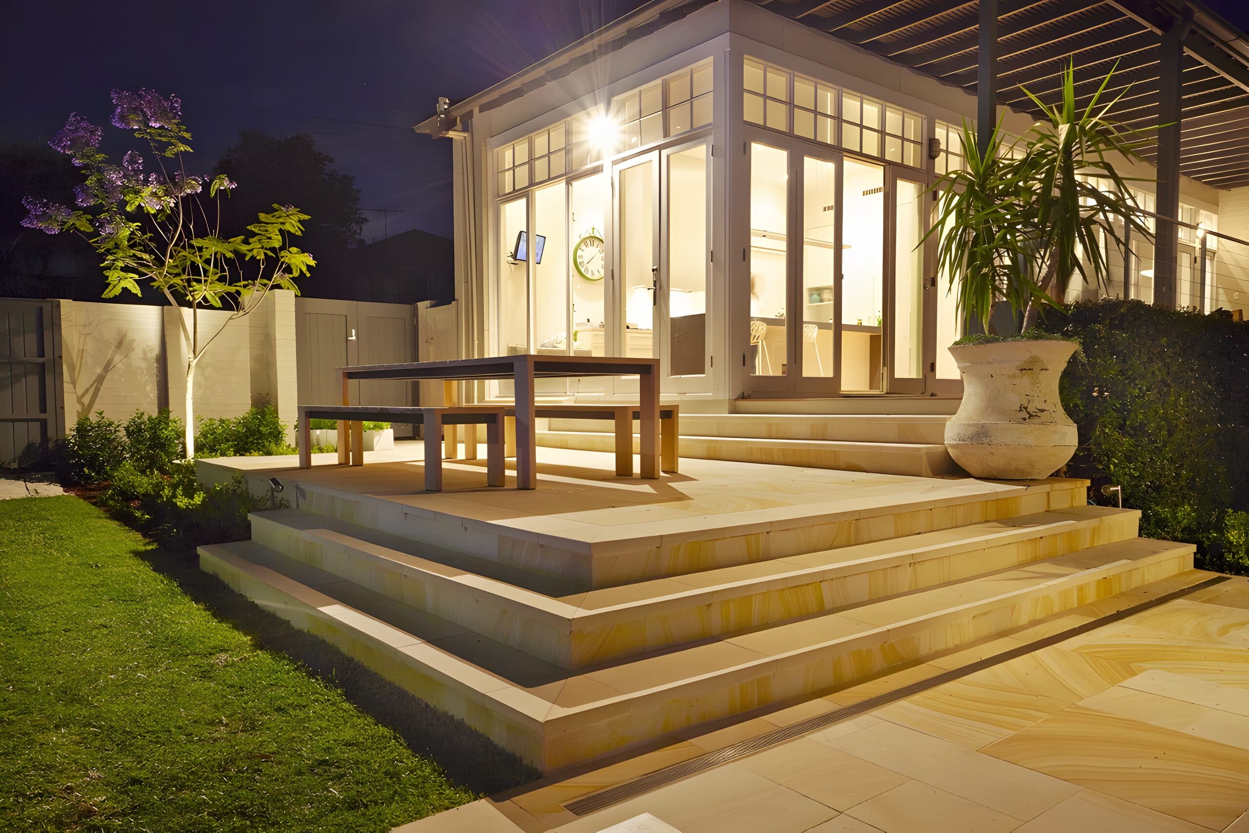 Illuminated garden room glowing at night above a sandstone platform set with a timber dining setting