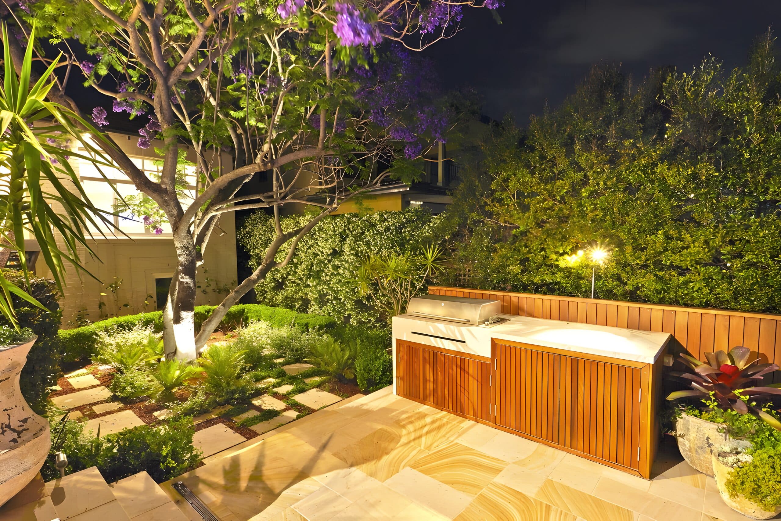 Outdoor kitchen and uplit jacaranda at night beside a chequerboard path through lush planting