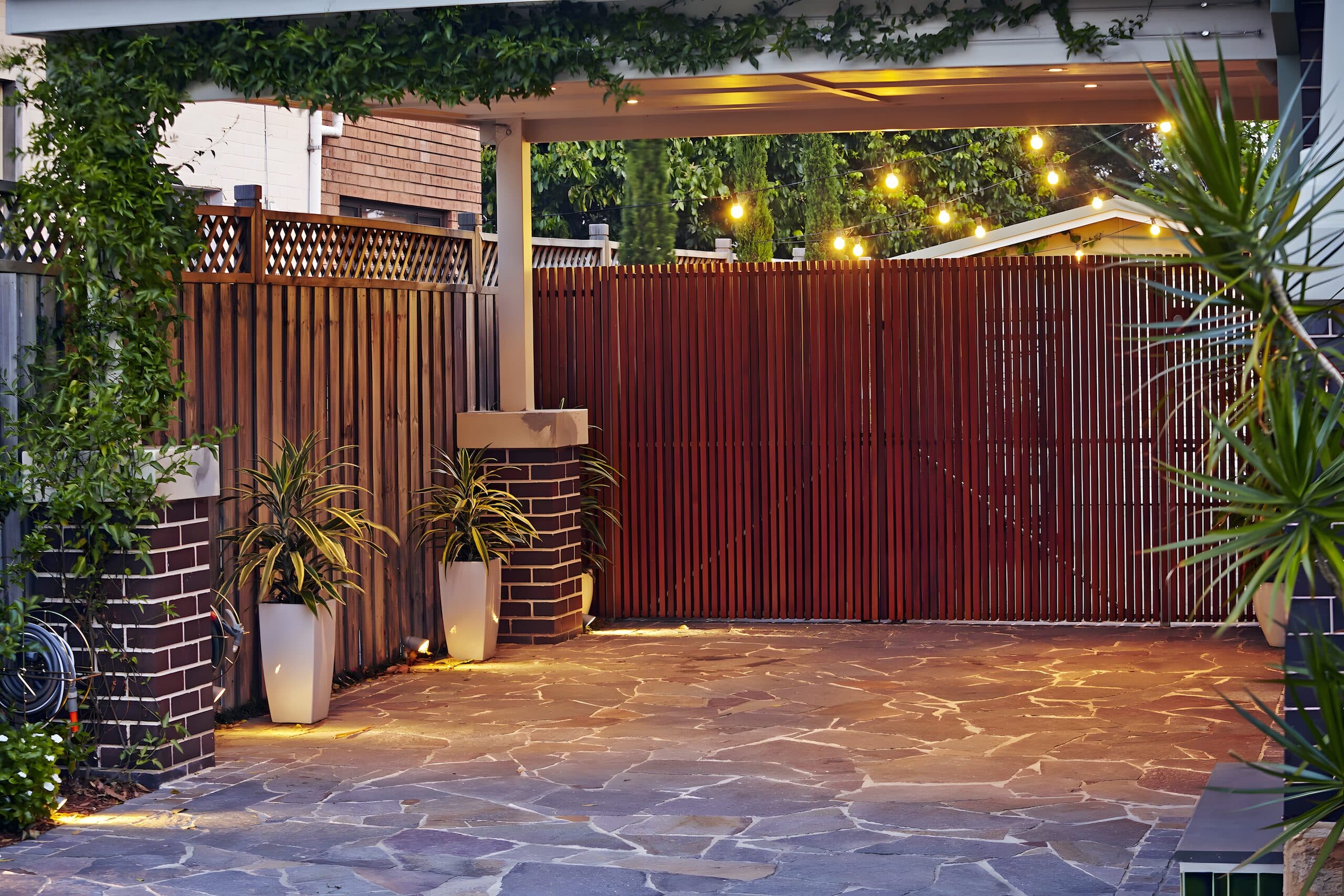 Festoon-lit carport with timber slat gates and potted cordylines over crazy paving at dusk