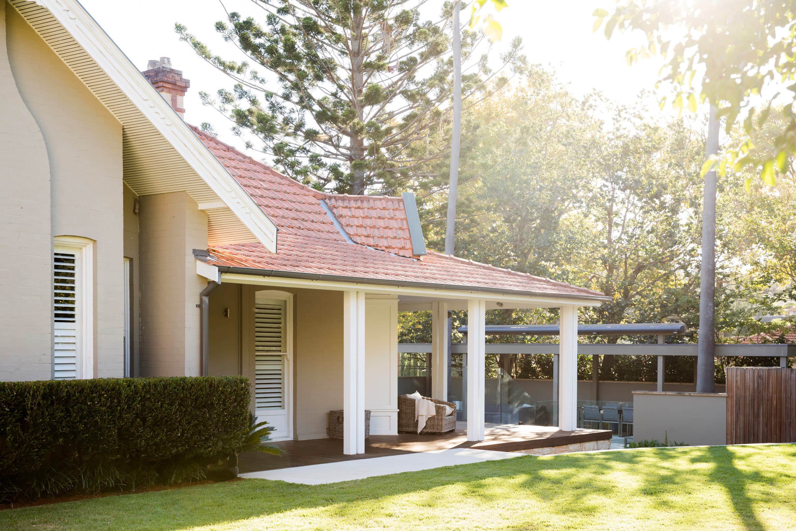 Sunlit cream brick home with terracotta roof and covered verandah beside a clipped hedge and lawn