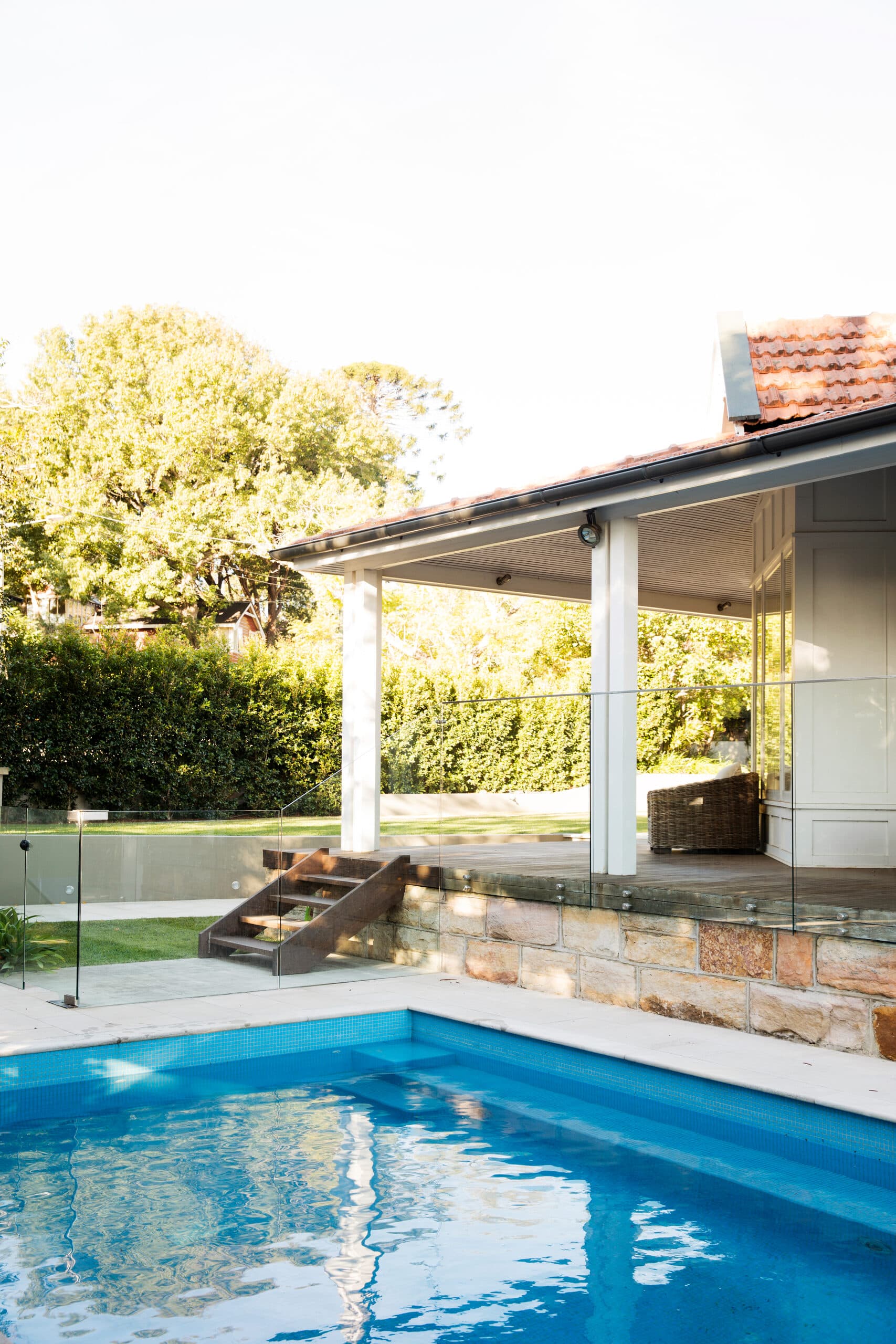 Blue tiled pool with frameless glass fencing and sandstone retaining wall below a timber stair and verandah