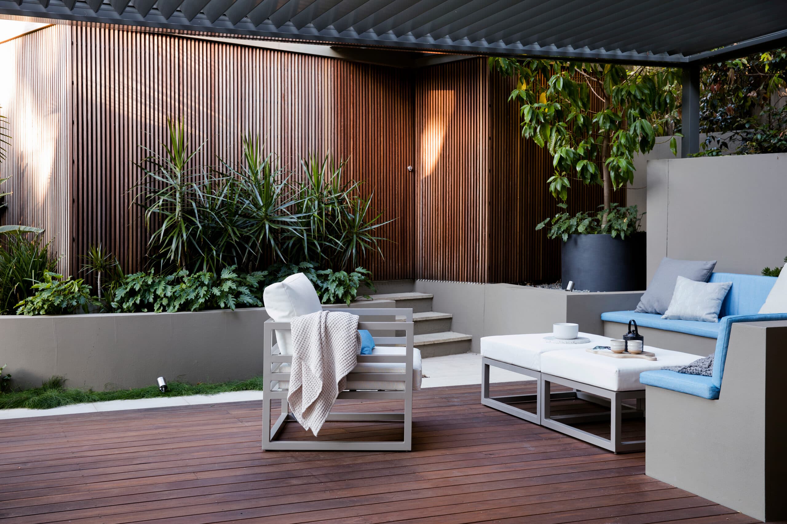 Covered timber deck with grey armchairs and a built-in bench of blue cushions against a timber slat screen and dracaenas