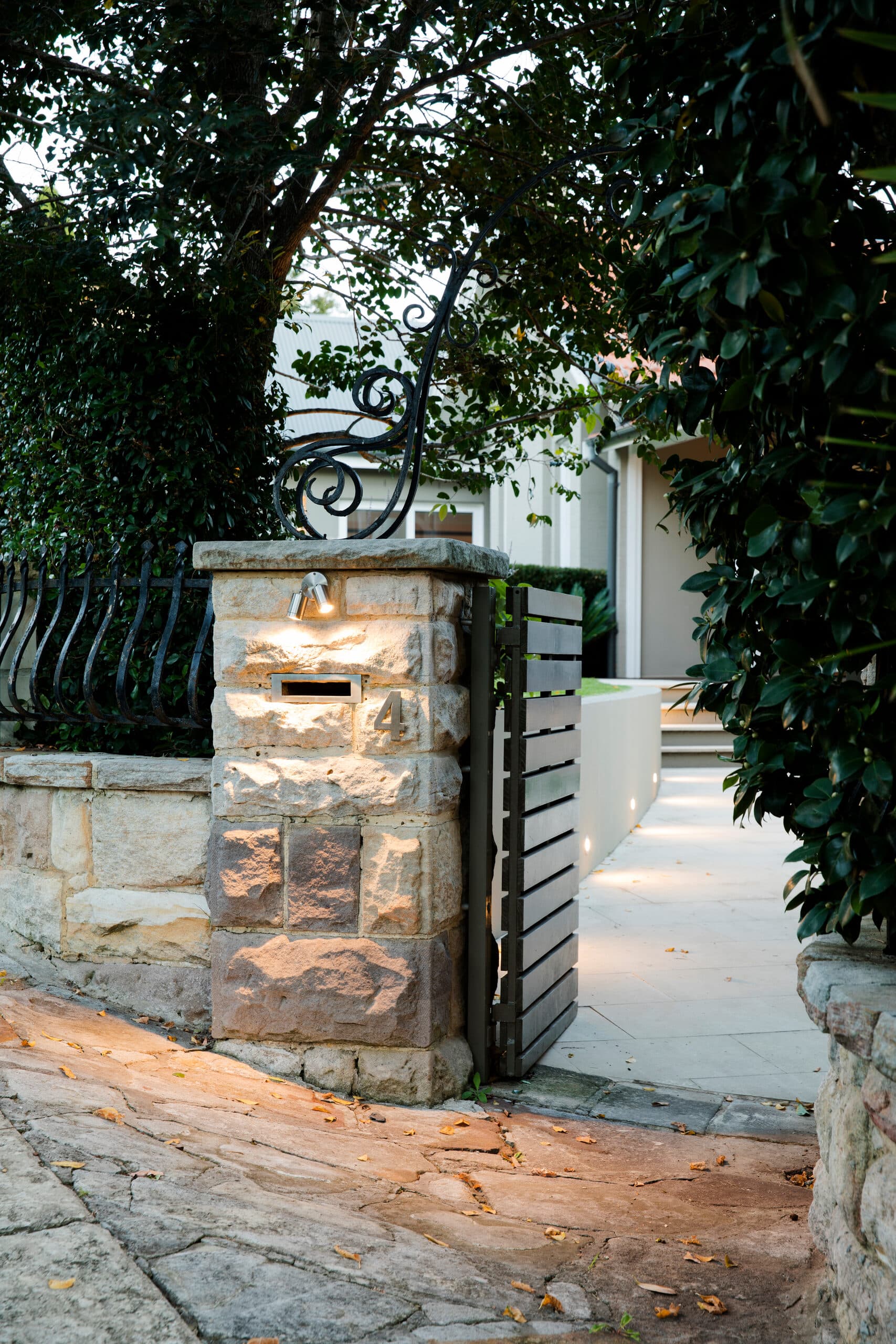 Sandstone gate pier with a numbered letterbox and slatted timber gate lit along the entry path at dusk