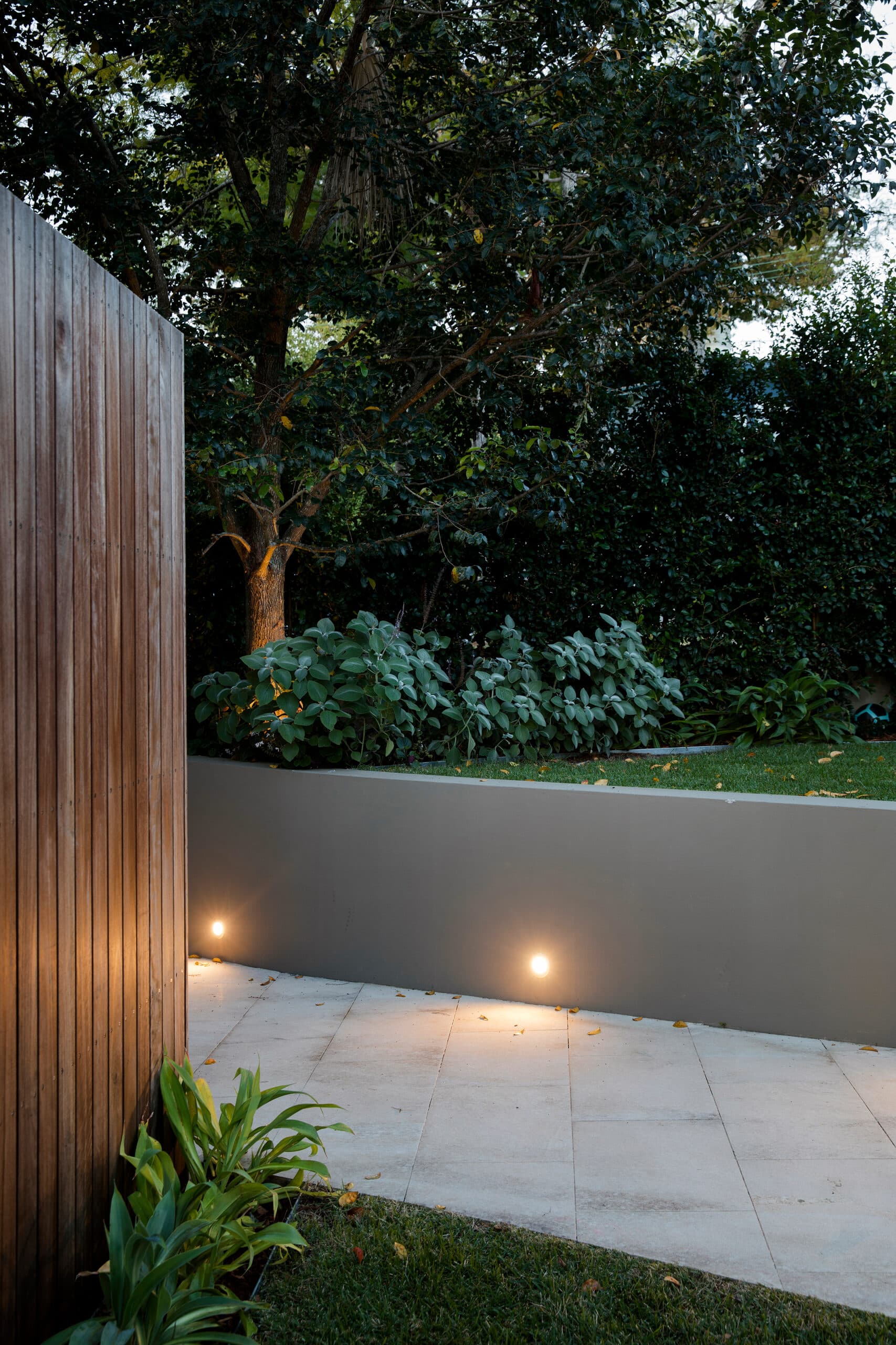 Recessed wall lights washing sandstone paving at dusk beside a timber screen and leafy boundary