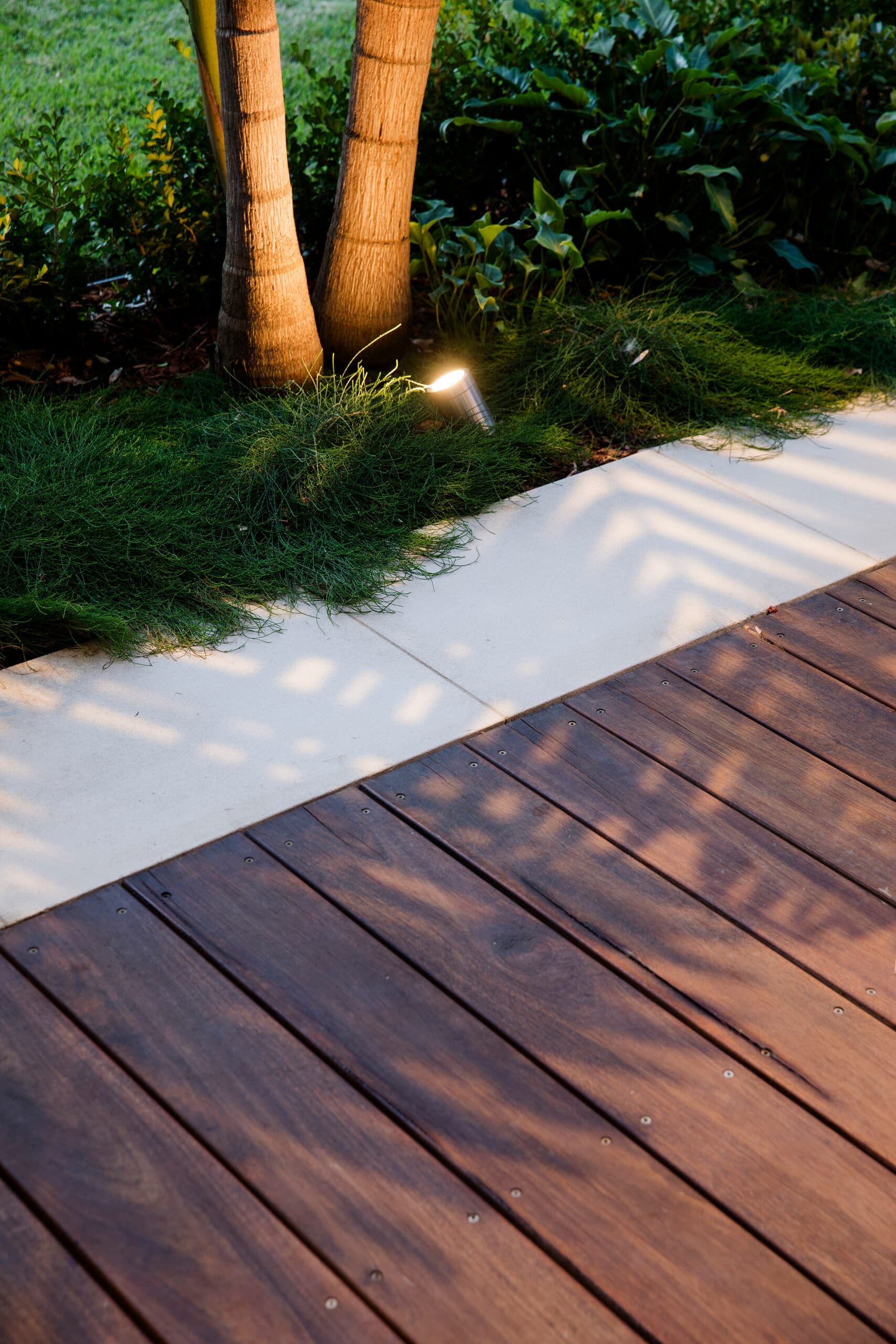 Detail of rich timber decking meeting a sandstone paver strip with mondo grass and a lit palm trunk