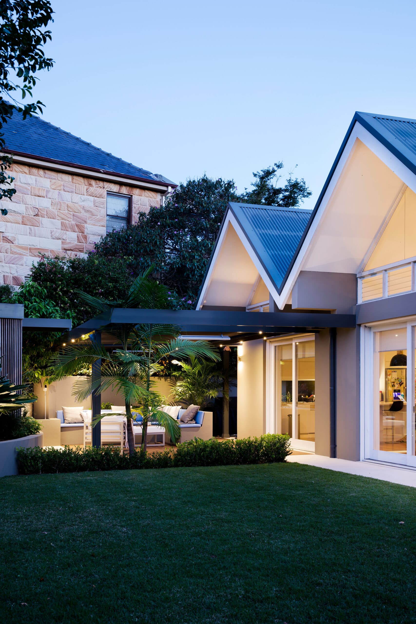Gabled home glowing at twilight with a lit lounge under the pergola and lawn framed by sandstone