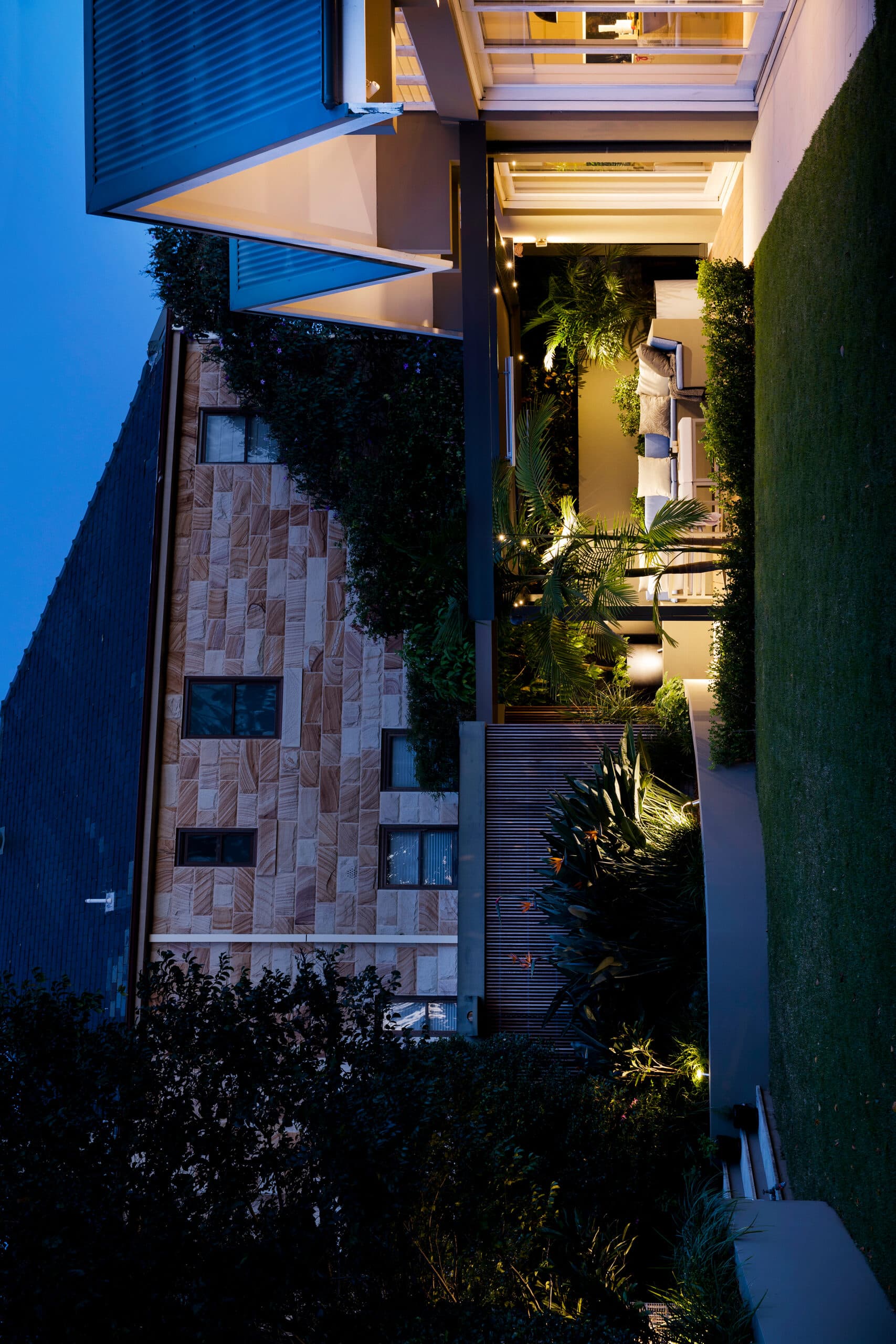 Dusk view across lawn to a softly lit outdoor lounge below the sandstone home with birds of paradise glowing