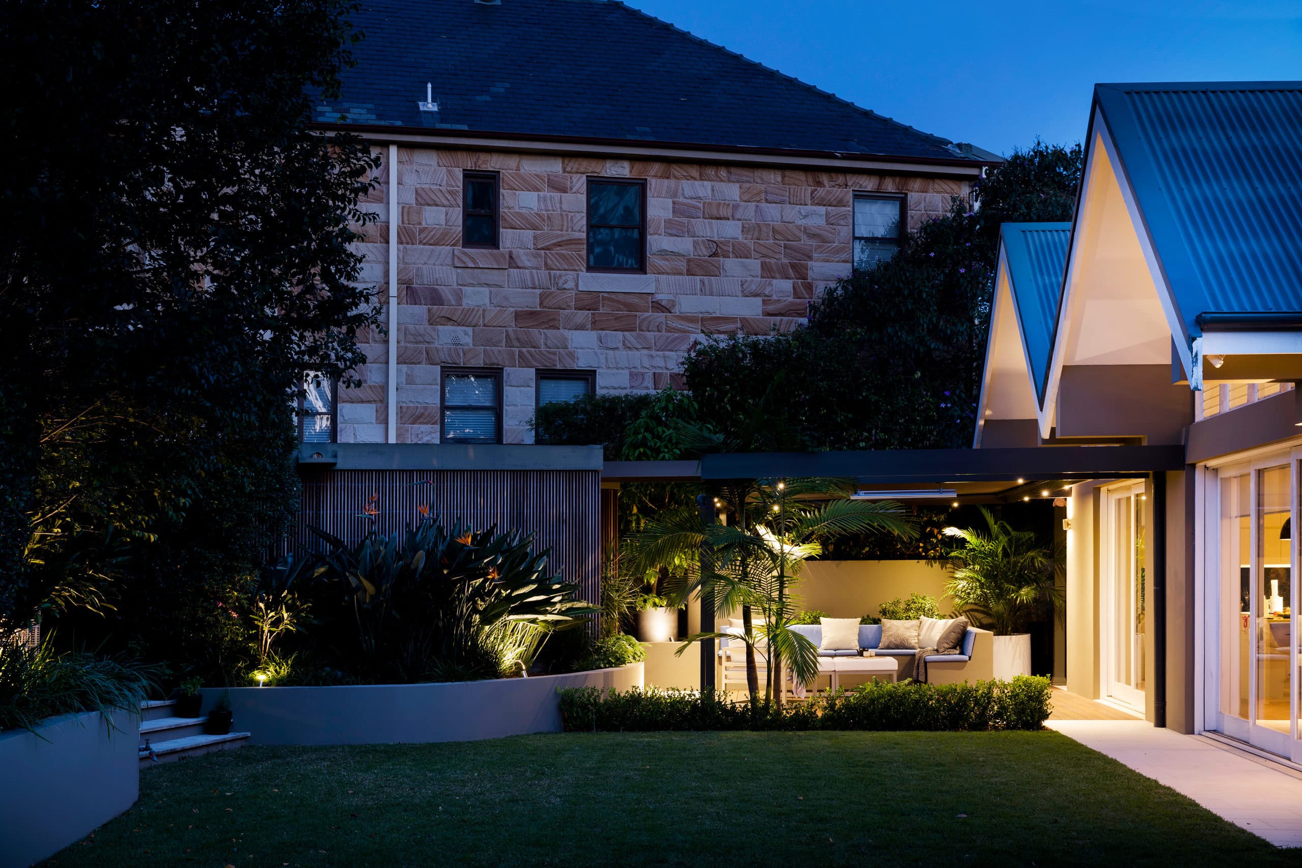 Dusk view across lawn to a softly lit outdoor lounge below the sandstone home with birds of paradise glowing