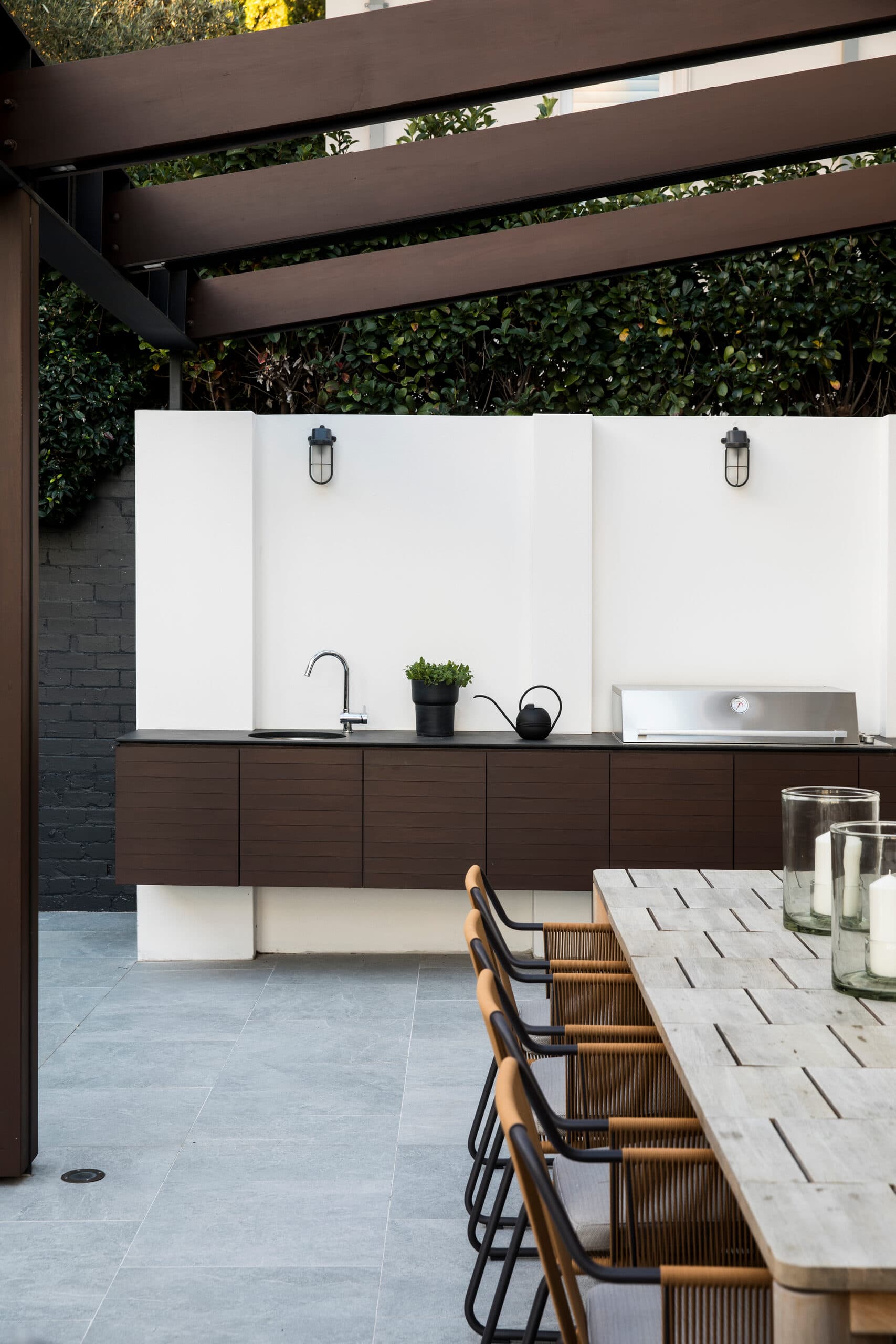 Covered outdoor kitchen with dark timber cabinetry, sink and barbecue beside a long dining table