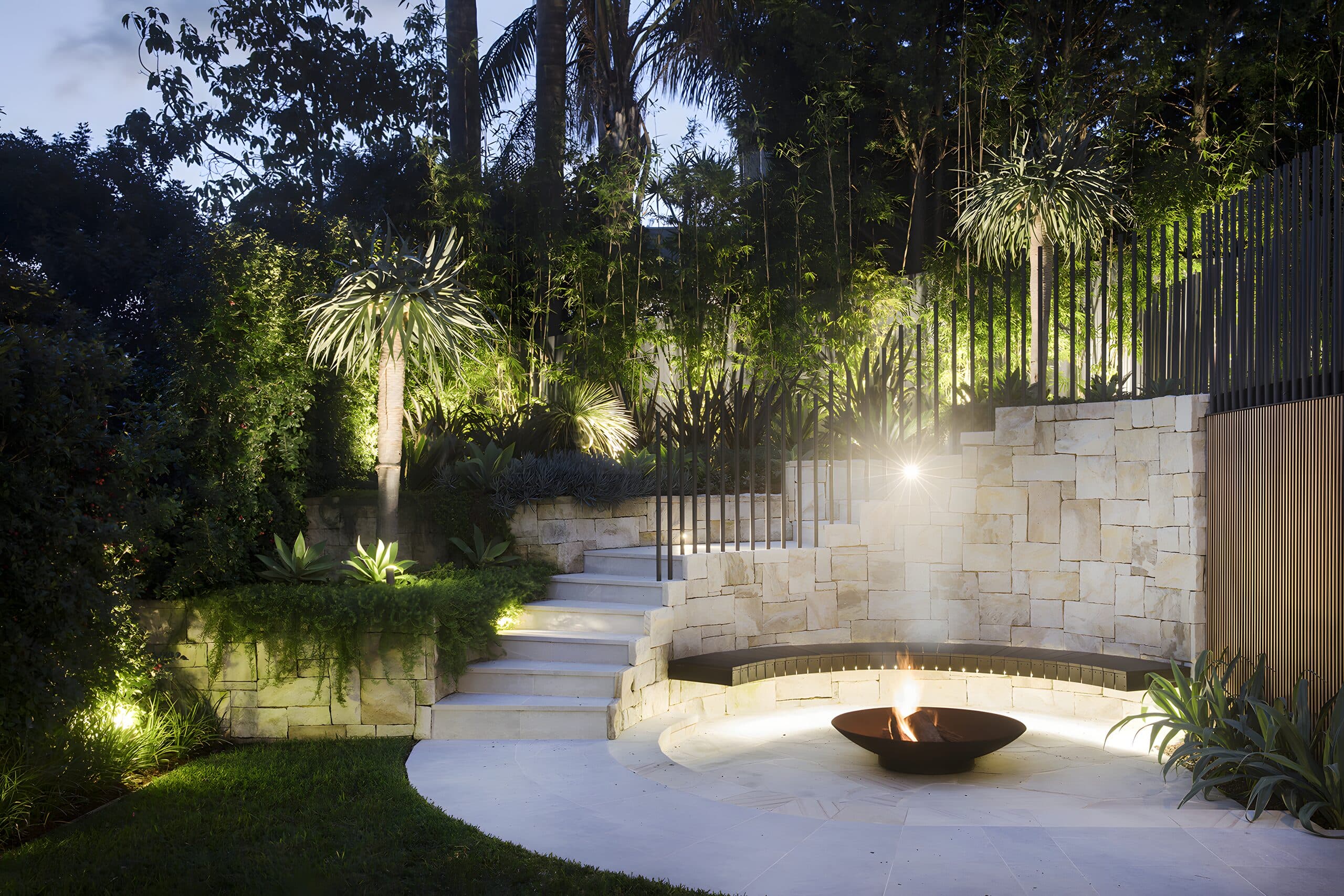 Firepit courtyard lit at night with uplit palms, sandstone wall and curved bench seat