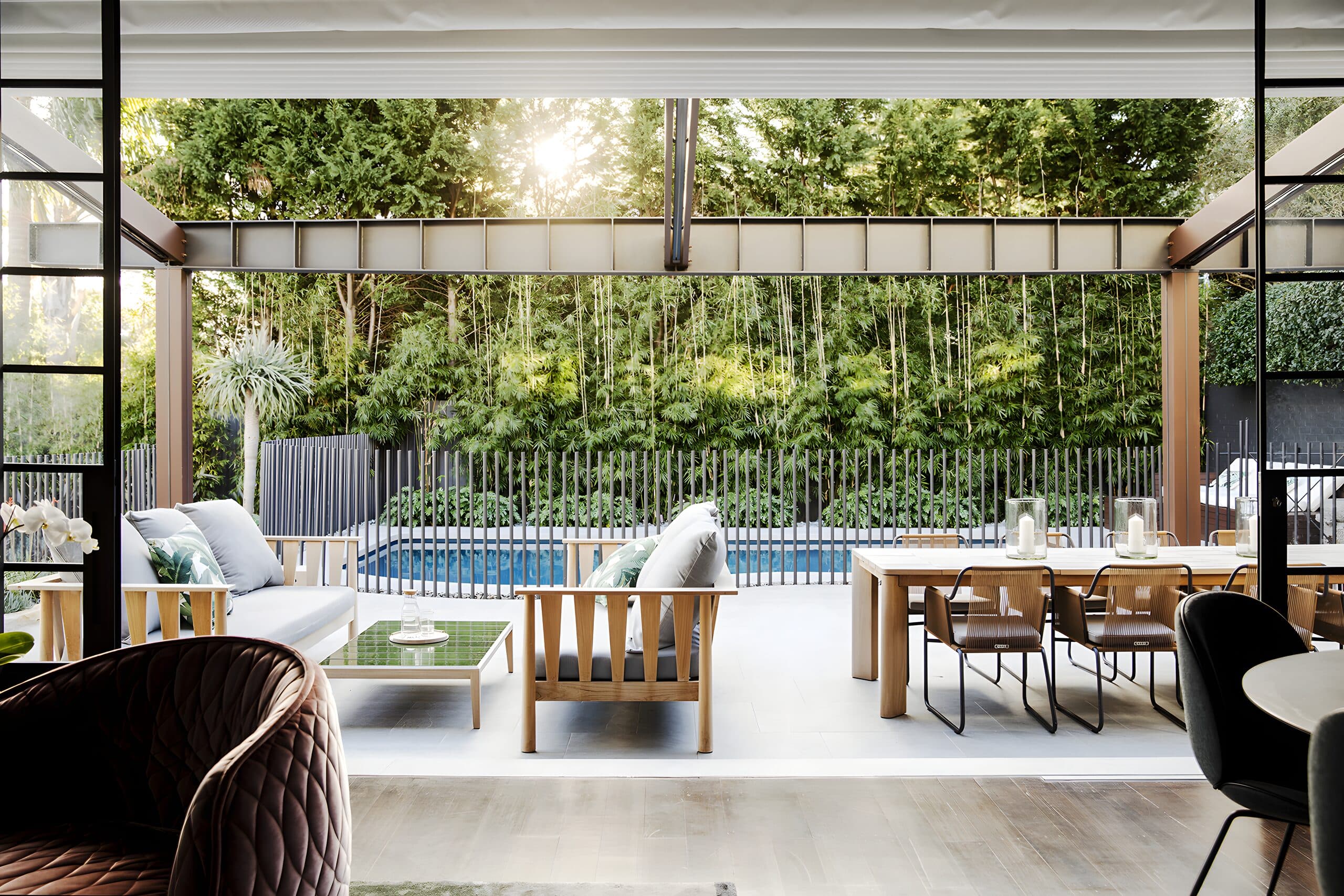 View from a covered lounge across timber furniture to the pool and tall bamboo screen