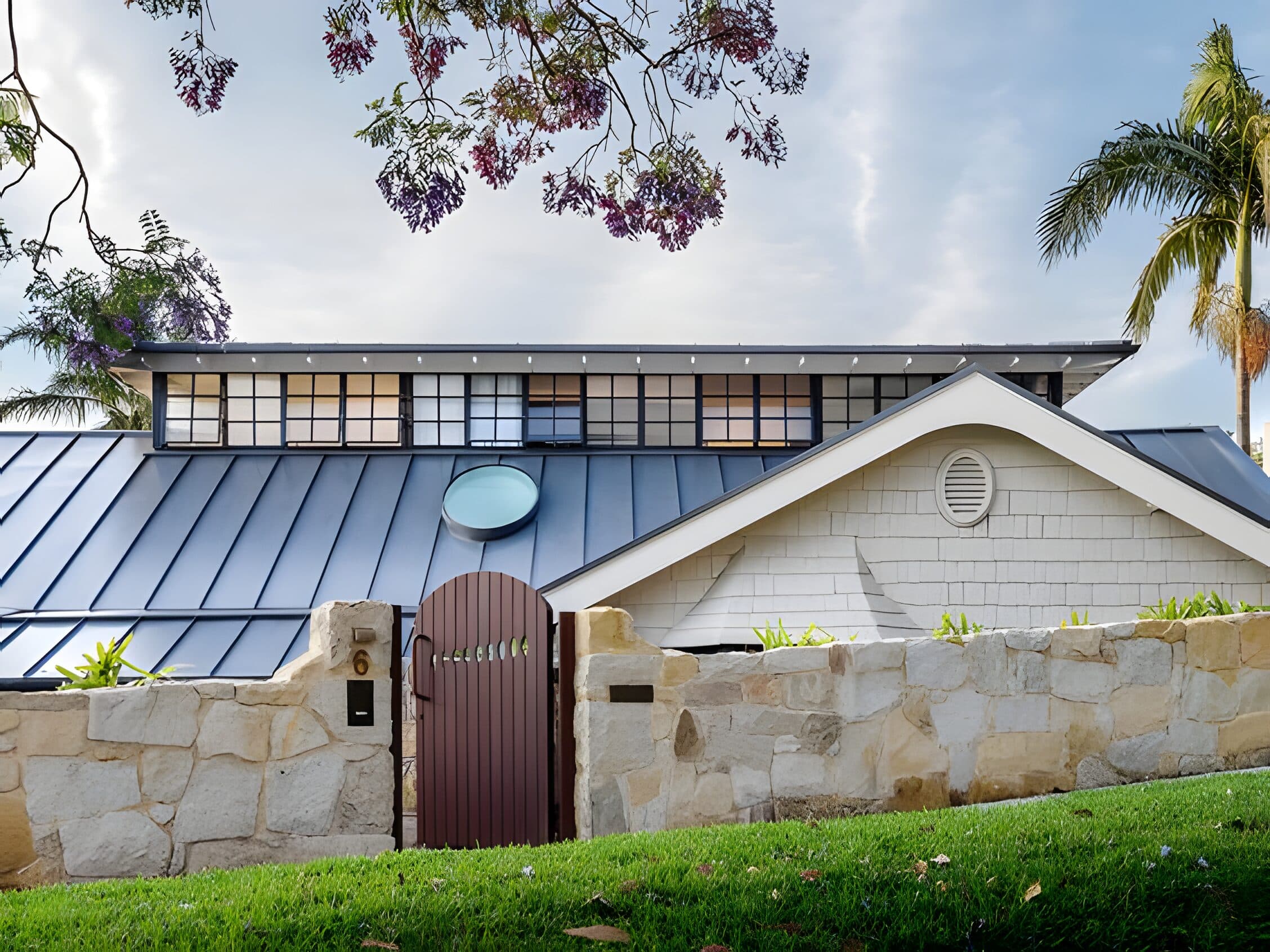 Street view of the house with metal roof and sandstone wall framed by flowering jacaranda
