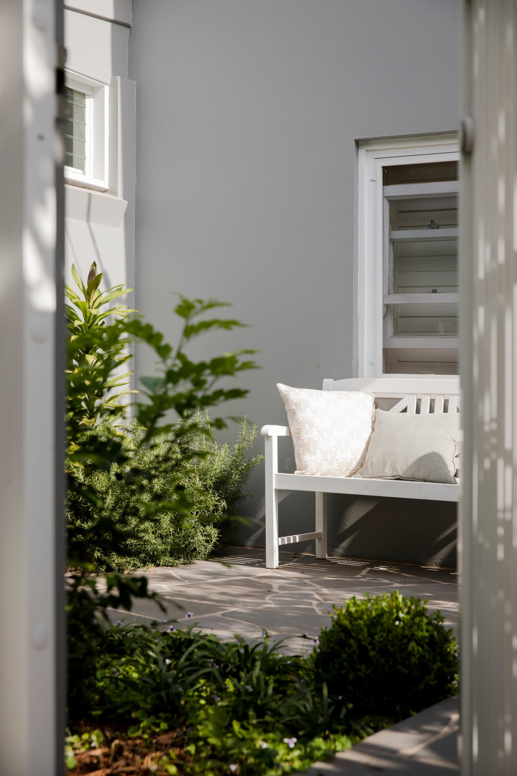 White timber bench with cushions in a sunlit courtyard framed by shrubs and crazy paving