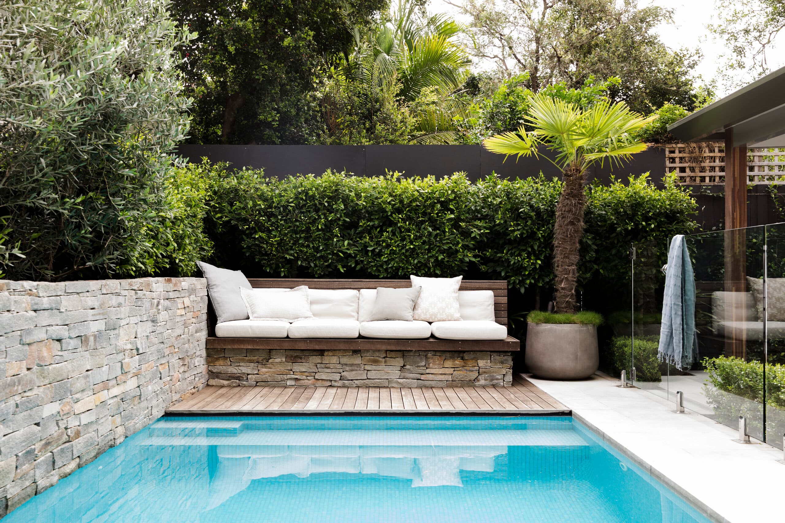 Stone-clad daybed with white cushions on timber decking beside the pool and glass fence