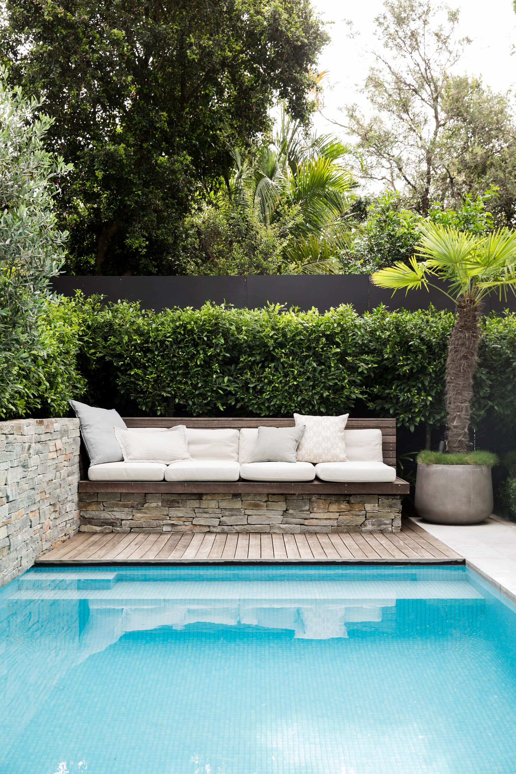 Built-in timber daybed on a stone base beside the pool below a tall clipped hedge