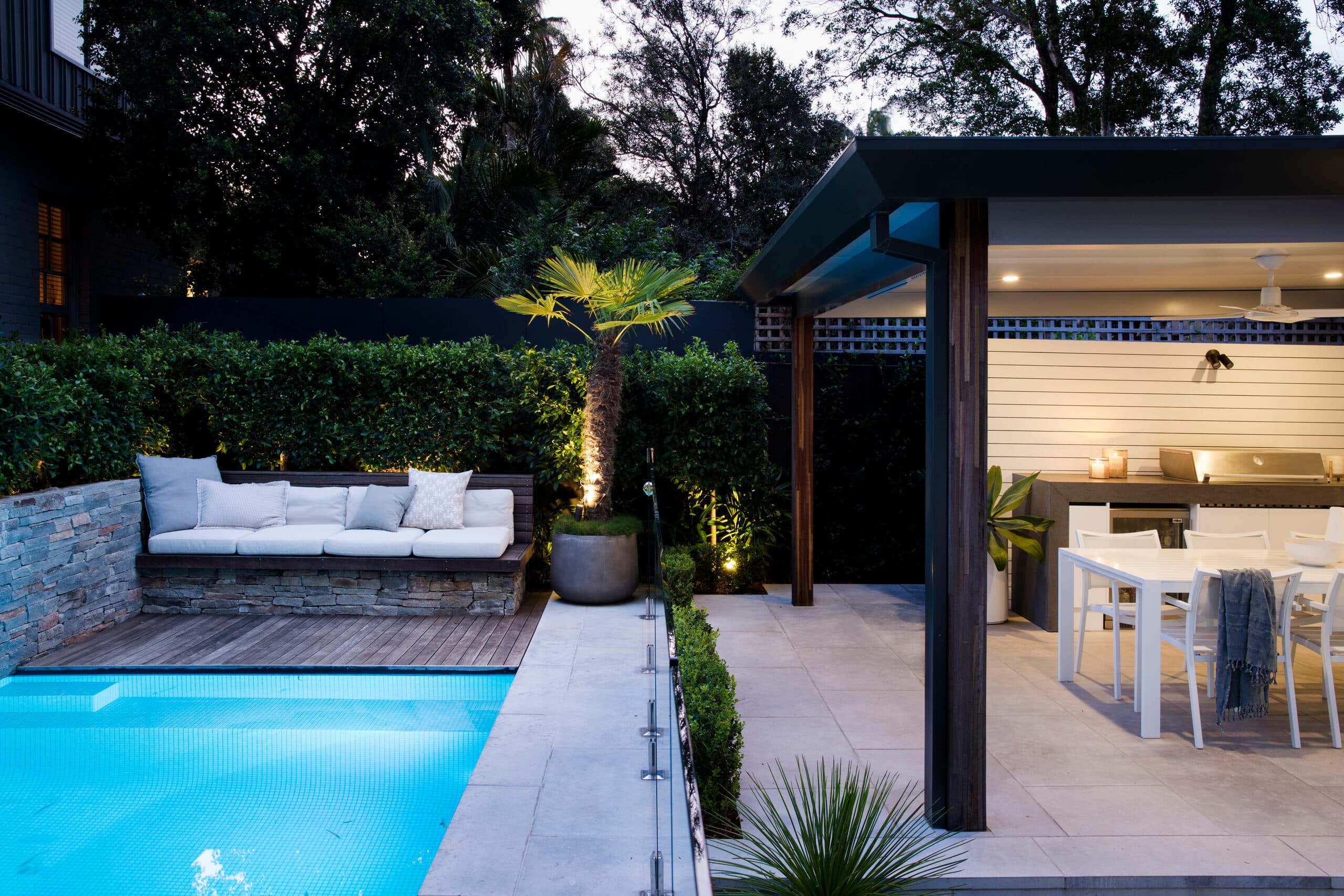 Dusk view of the lit pool and daybed with an uplit palm beside the alfresco kitchen