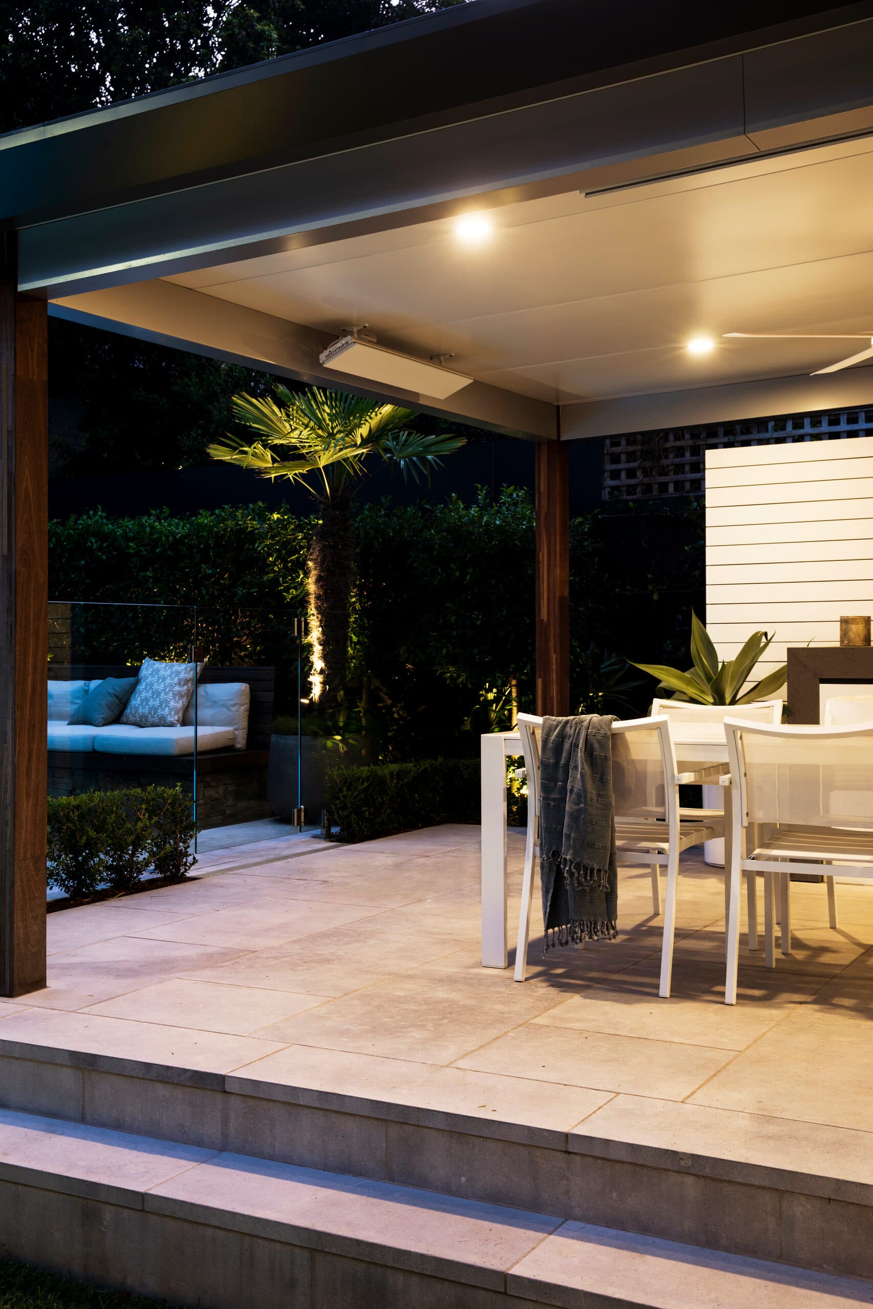Alfresco at night with warm downlights white dining and an uplit palm beyond the glass fence