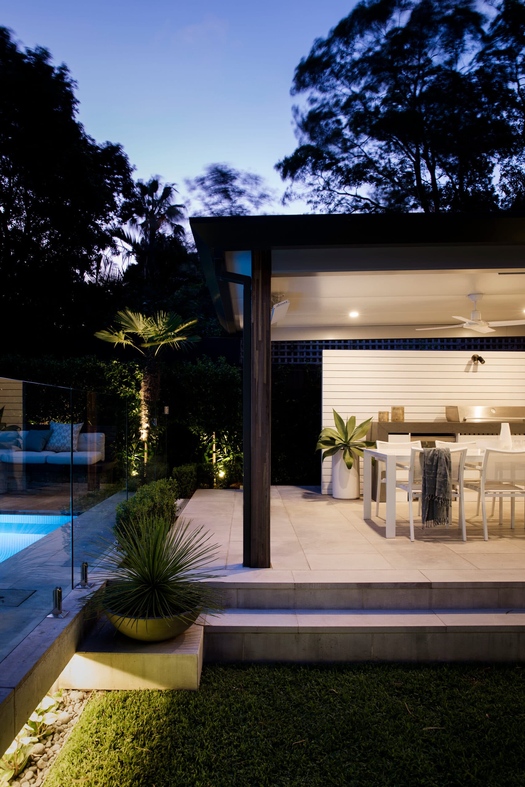 Blue-hour view of the lit alfresco pavilion with dining and an uplit palm beside the pool