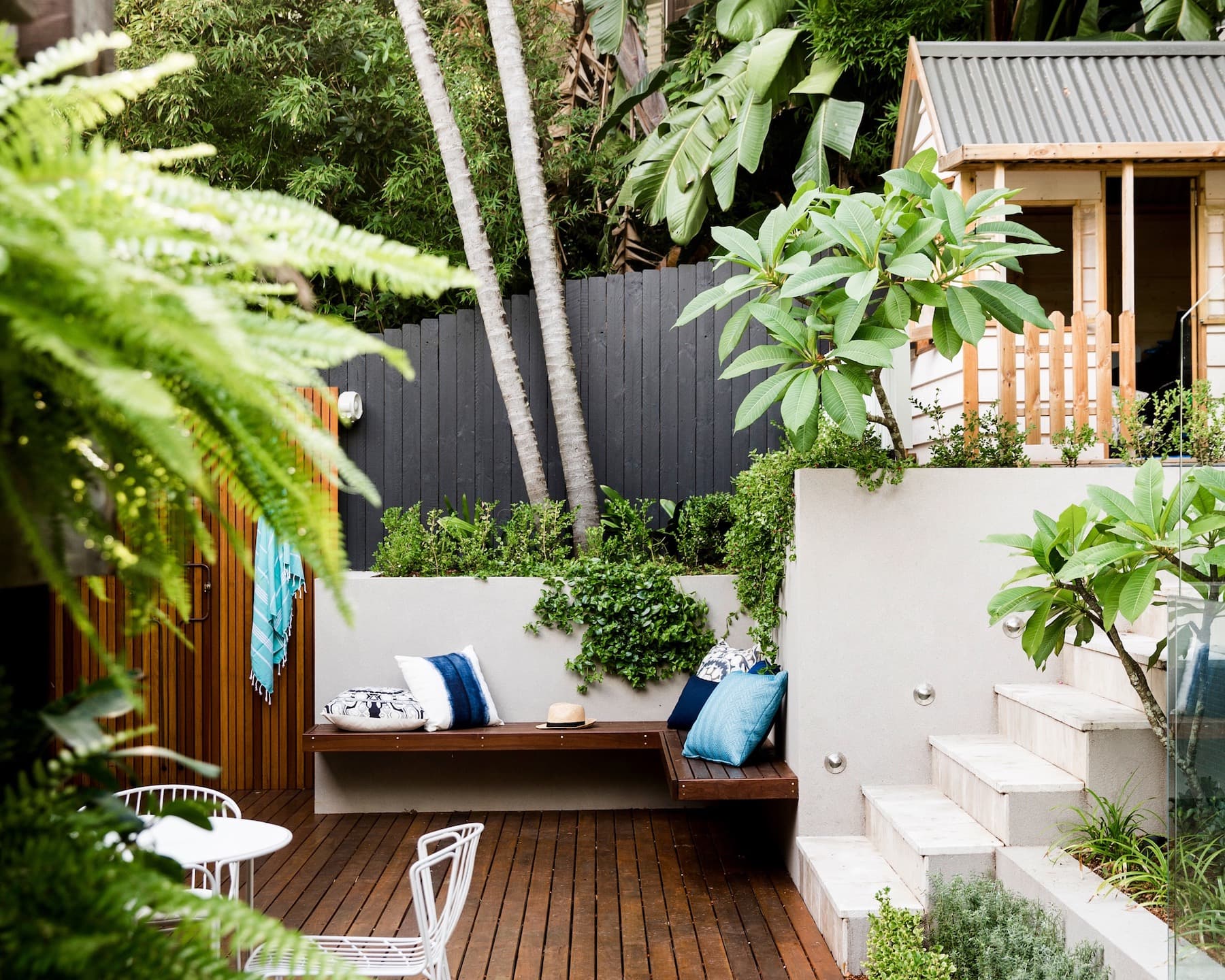Floating timber bench with blue cushions tucked under palms beside sandstone steps and a cubby roof