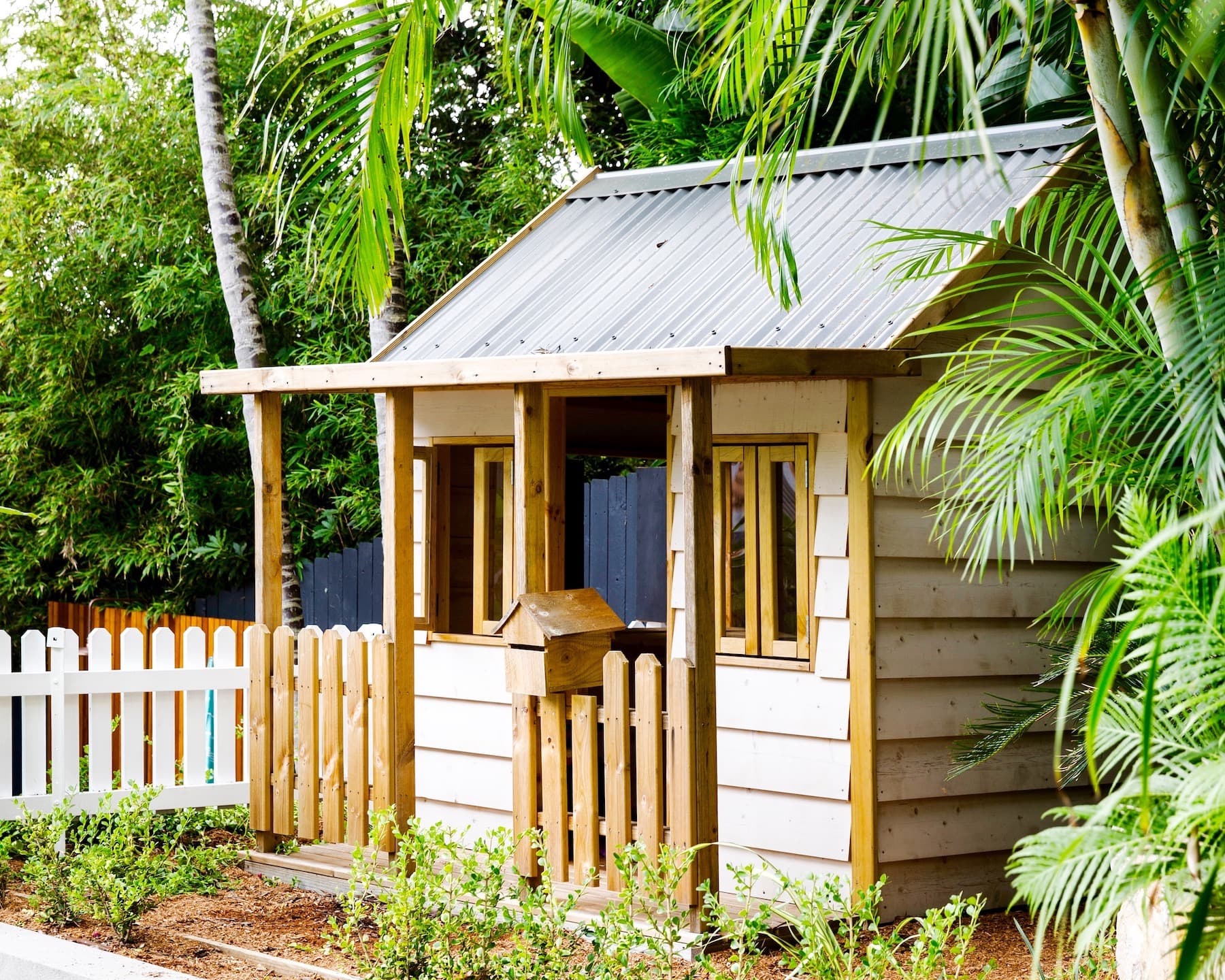 Timber cubby house with a corrugated metal roof beside a white picket fence and palms