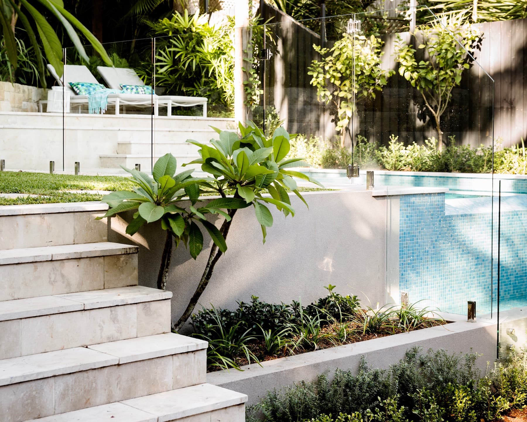 Sunlit sandstone steps and frangipani beside a glass fenced mosaic pool with loungers on the upper terrace